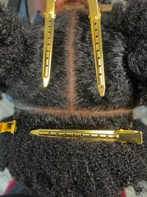 Close-up of a person's head with curly black hair sectioned and clipped with gold hair clips, revealing the scalp and parting lines.