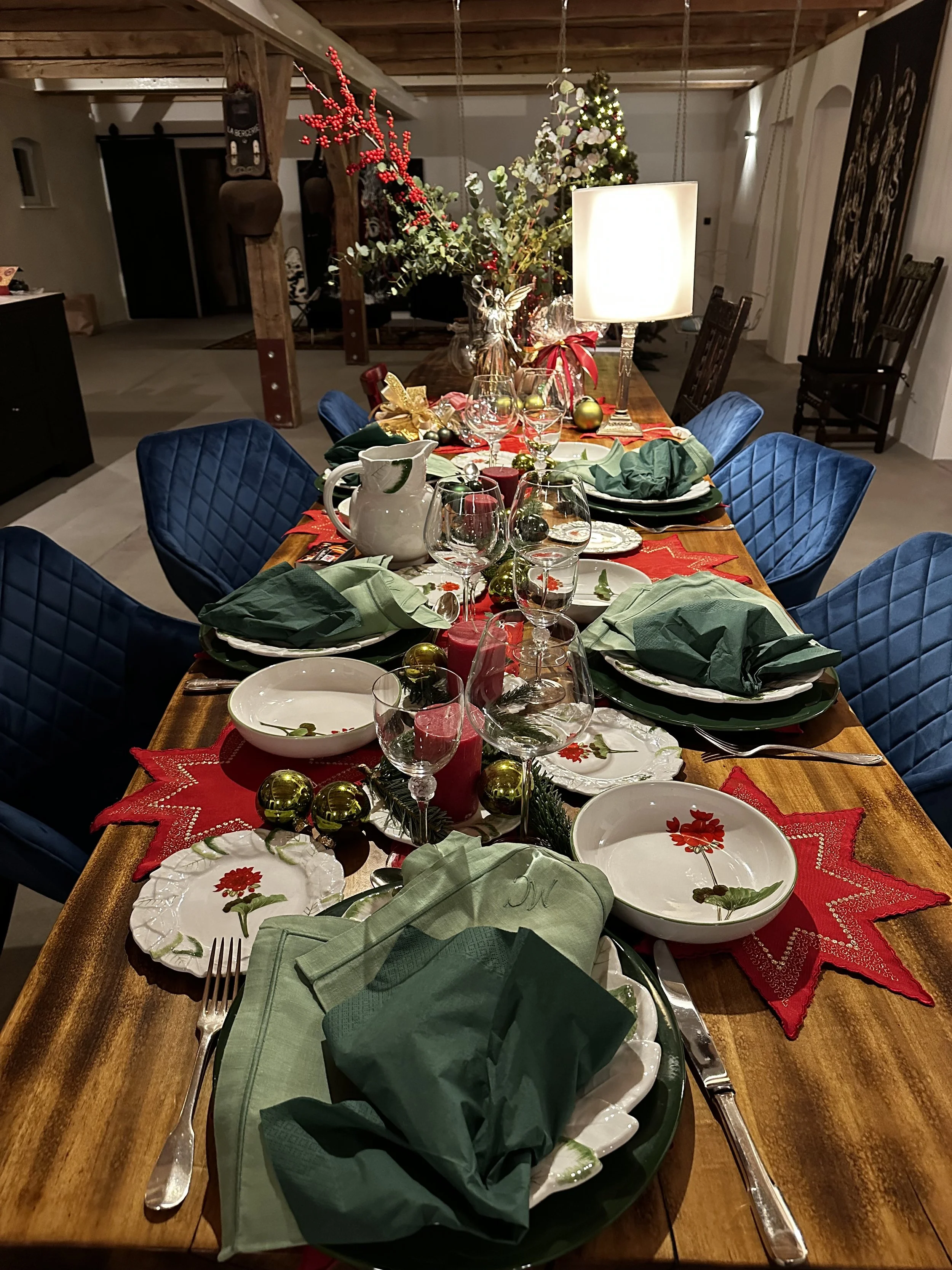 Ein festlich gedeckter Esstisch mit Weihnachtsdekoration, grünem und rotem Design, Kerzen und Geschirr, in einem gemütlichen Raum mit Weihnachtsbaum im Hintergrund.