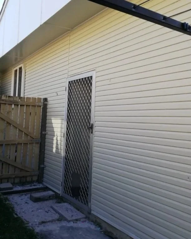 Exterior view of a house showing a beige vinyl siding wall, a door with a metal security screen, a wooden fence on the left, and part of the roofline at the top.