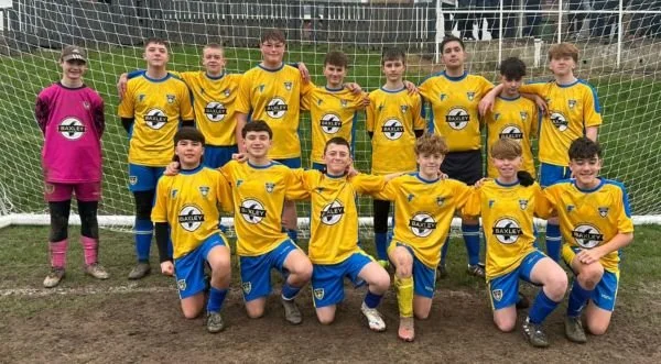 A youth soccer team wearing yellow jerseys and blue shorts, posing on a soccer field in front of a goal, with 11 players and a goalie in a pink jersey.