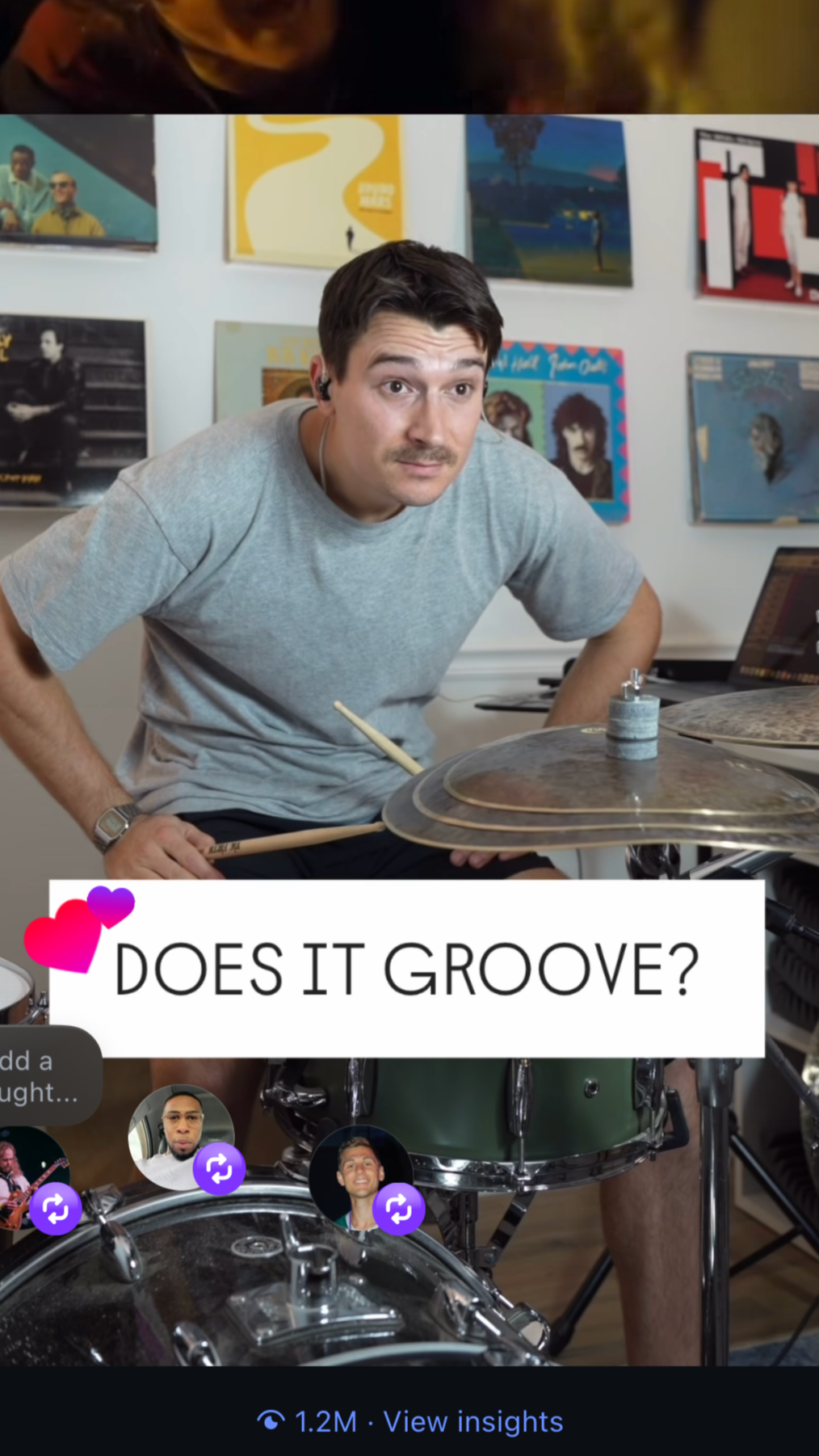 A man with dark hair and a mustache playing the drums in a room decorated with colorful album covers on the wall. He is wearing a gray t-shirt, earphones, and a wristwatch, and is looking at the camera. There is a text overlay that says 'DOES IT GROOVE?' with heart icons.