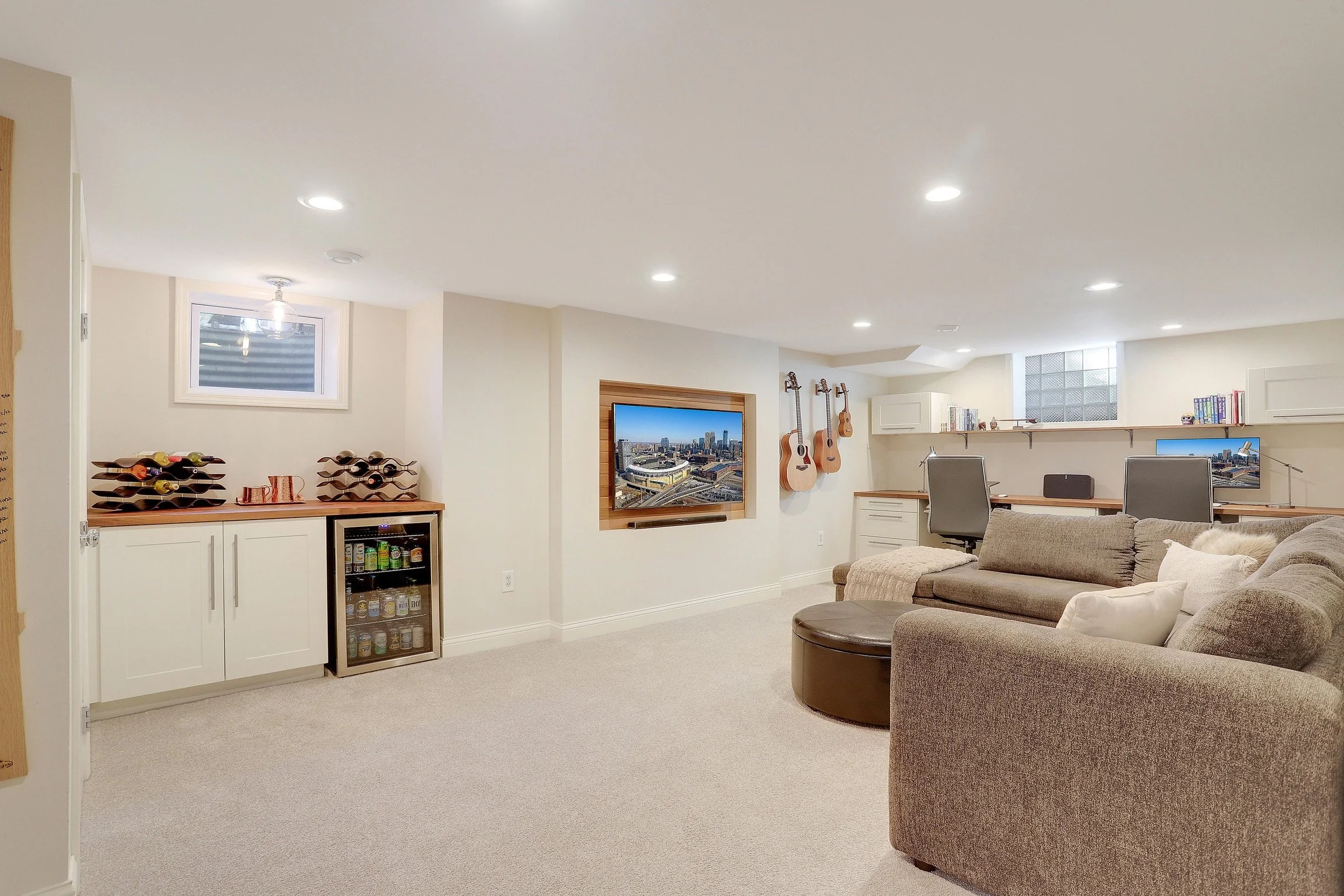 Basement entertainment room with a sectional sofa, wall-mounted TV, guitars on the wall, and a workspace area with two desks and chairs.