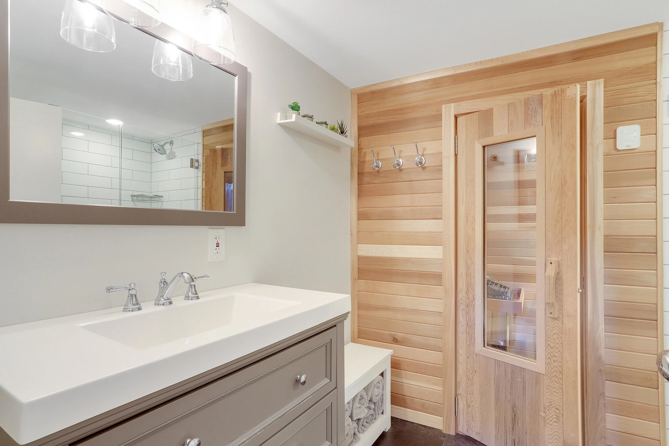 A bathroom with a white sink vanity, a mirror, and a shower visible in the mirror's reflection. The wall to the right is decorated with horizontal wood paneling, featuring hooks and a small windowed door leading to a sauna or steam room. There is a w
