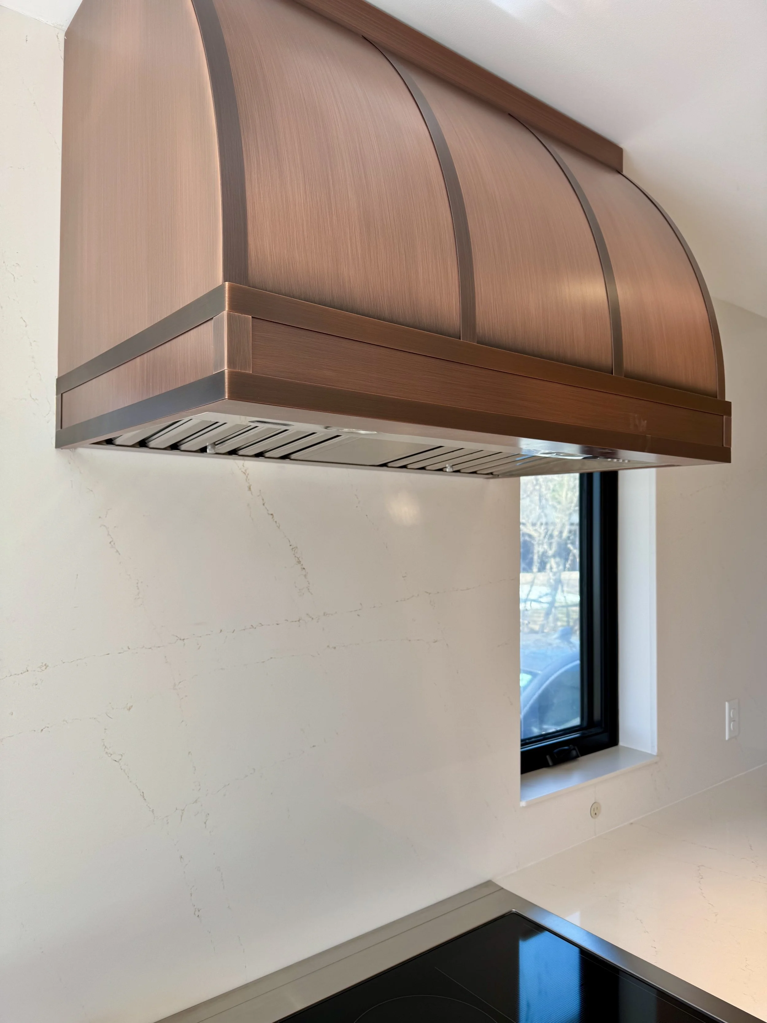 Copper-colored modern kitchen range hood above a black cooktop, with a window in the background showing a car outside.