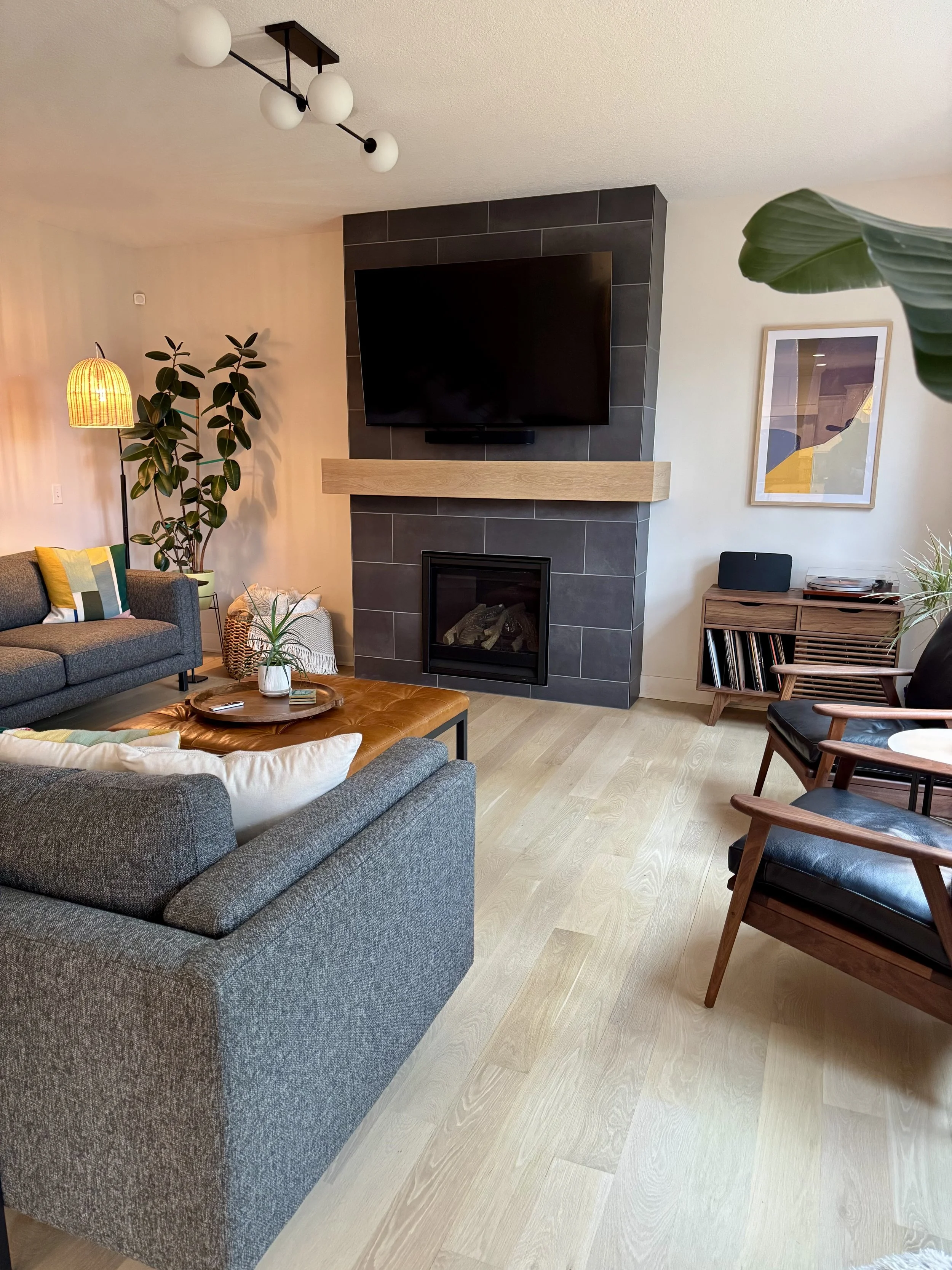 Living room with a gray sofa, orange leather ottoman, armchair with wooden frame and black cushion, fireplace, wall-mounted TV, potted plant, decorative items, artwork on the wall, and modern lighting fixtures.