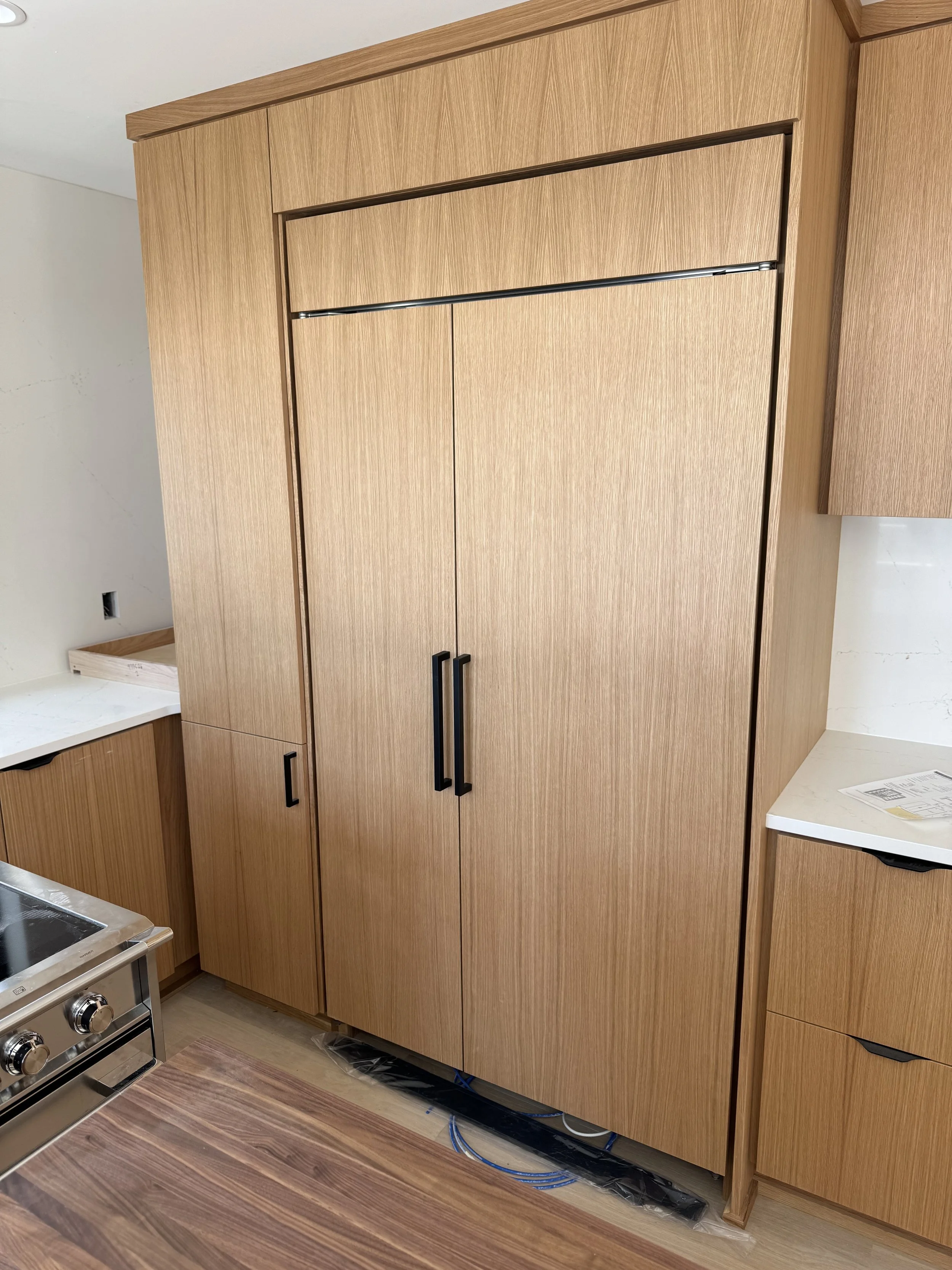 Wooden kitchen cabinets with black handles, adjacent to a stove and a white countertop, with wiring and a panel on the wall.