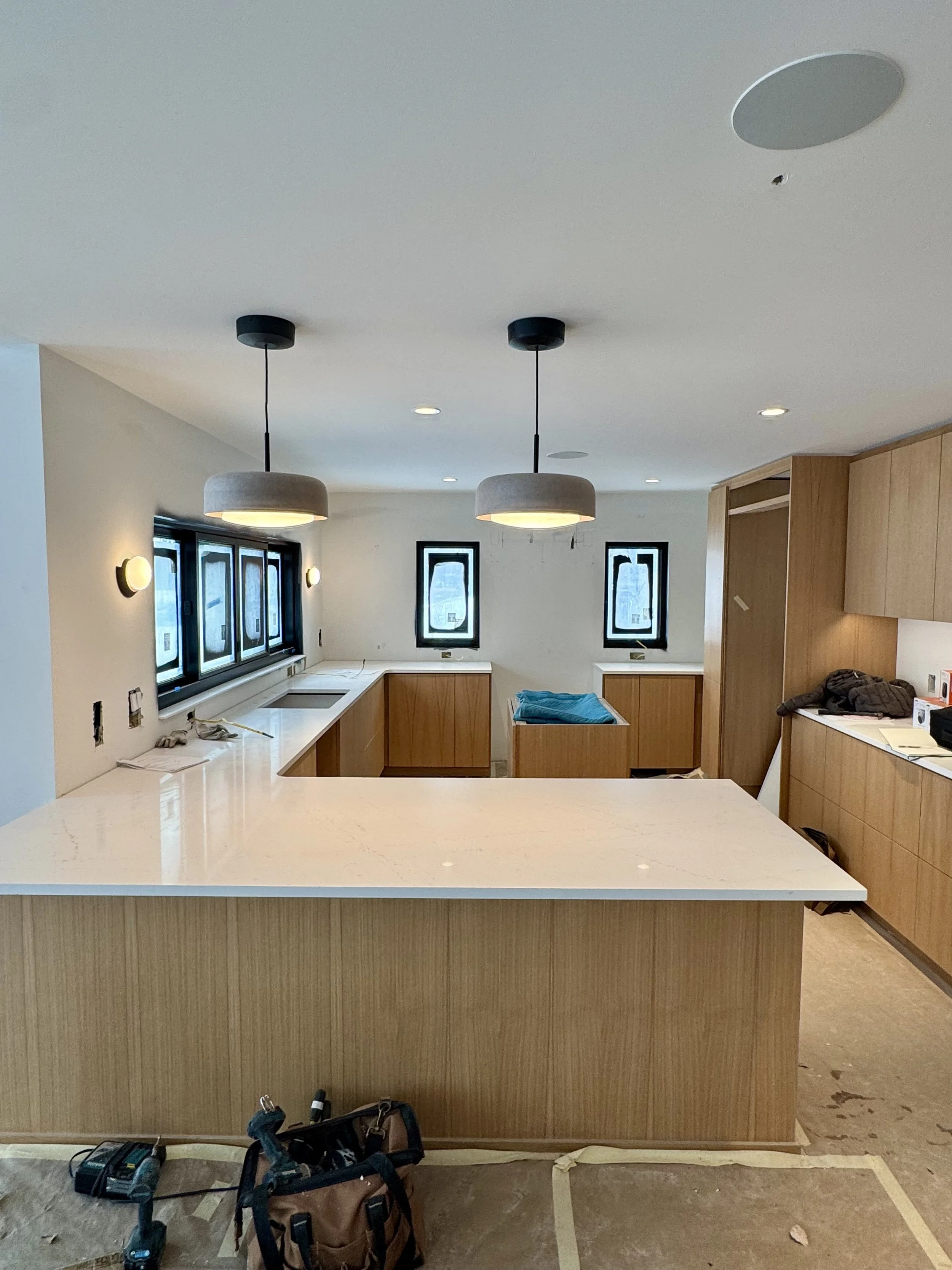 Modern kitchen under construction with wooden cabinets, white countertops, black framed windows, and hanging pendant lights.