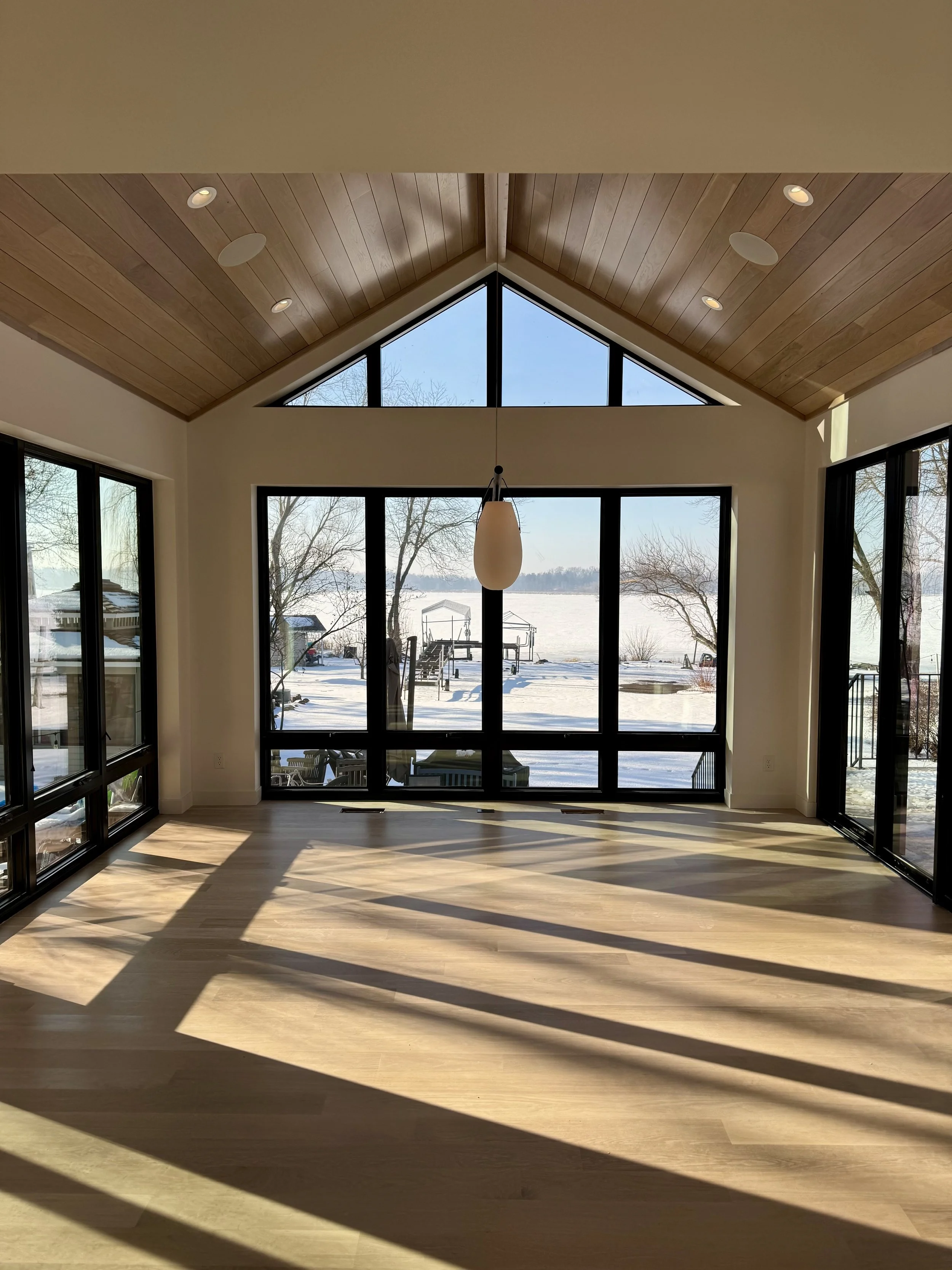 Empty room with large floor-to-ceiling windows overlooking a snowy outdoor landscape, wood-paneled vaulted ceiling, and sunlight casting shadows on the wooden floor.