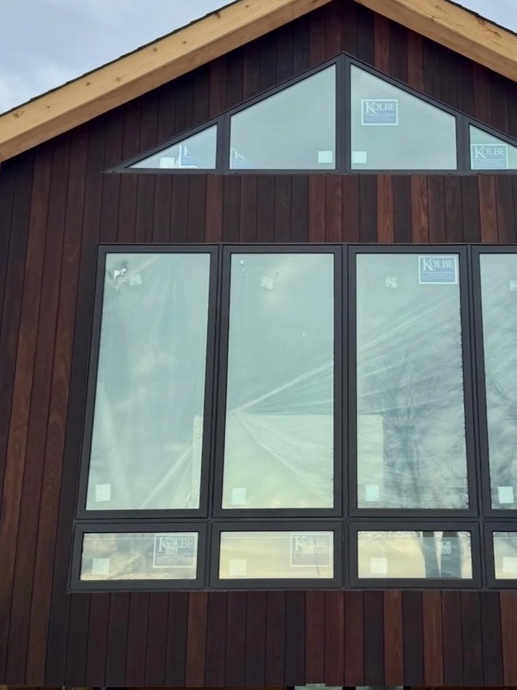 The image shows a modern wooden house exterior with large glass windows and a triangular top window.