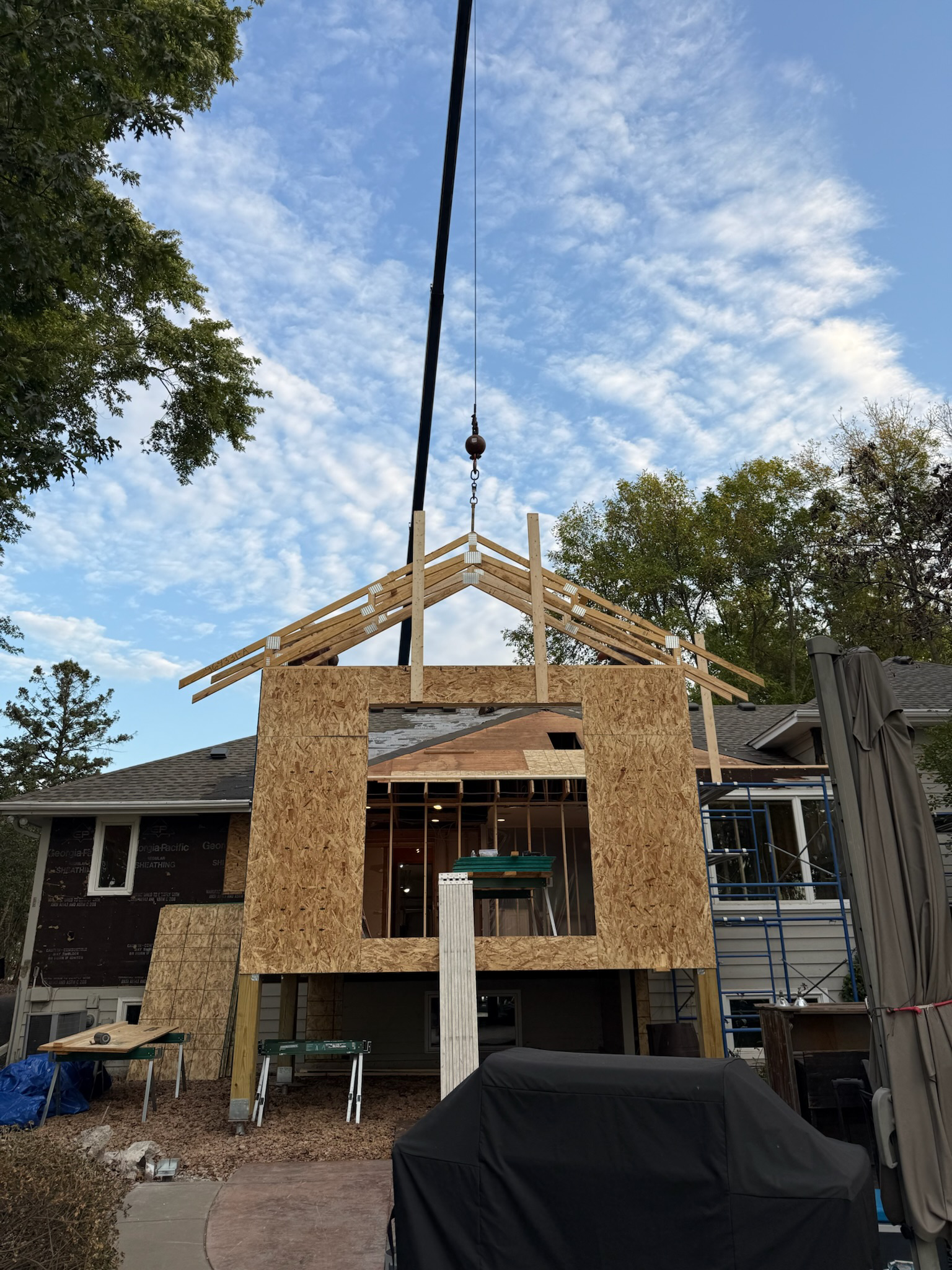 Construction of a house extension or new building with wooden framing and OSB sheathing, crane lifting roof trusses into place, in a residential neighborhood on a partly cloudy day.