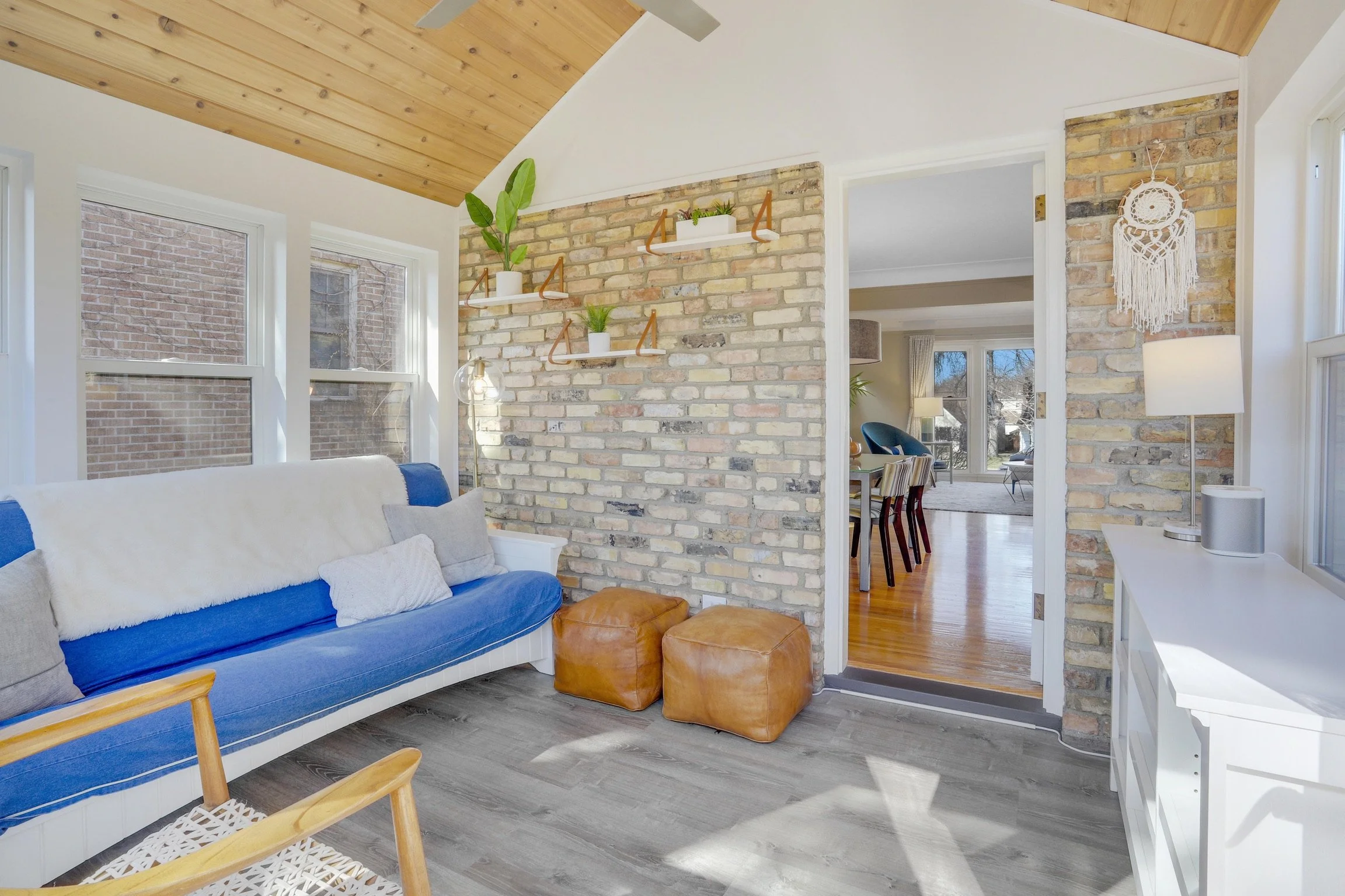 Inside a bright living room with white walls and large windows, a white couch with blue and gray pillows, two brown leather ottomans, a wooden chair, and a white sideboard. A brick accent wall features white floating shelves with plants, and a doorwa