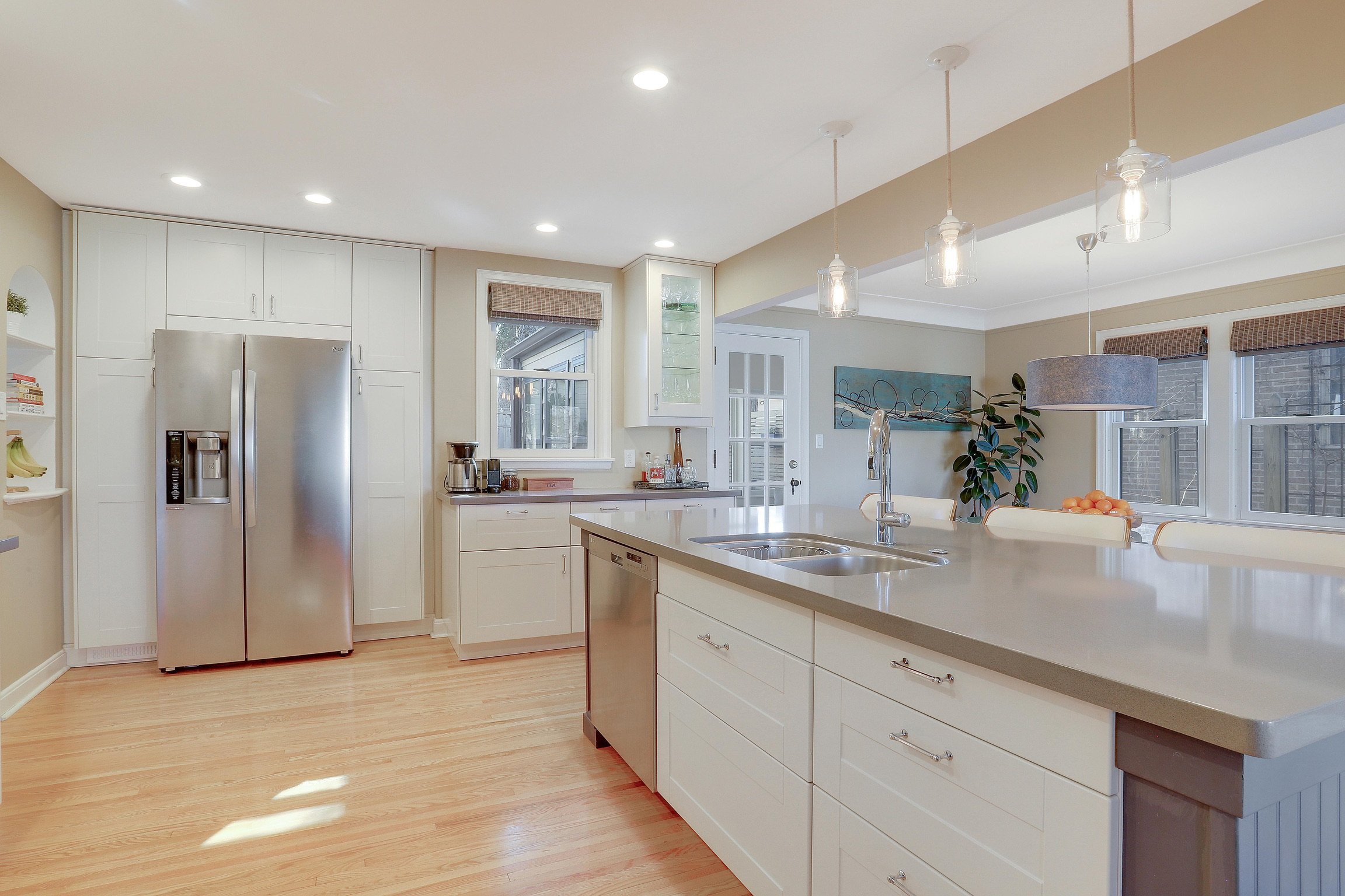 Modern kitchen with white cabinets, stainless steel refrigerator, island with sink, wooden flooring, and large windows.