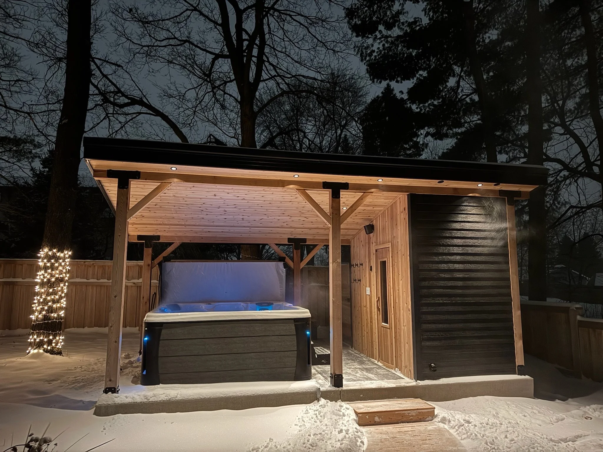 A modern outdoor hot tub with a wooden shelter in a snowy backyard at night, surrounded by trees and a wooden fence, with string lights wrapped around a tree on the left.