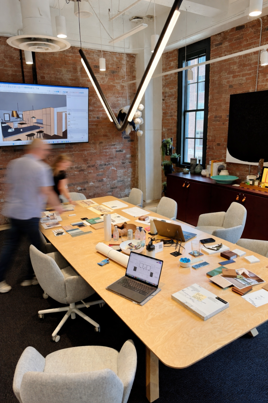 Modern brick-walled conference room with large wooden table, laptops, design materials, and a wall-mounted monitor displaying a room layout, with some people moving around.