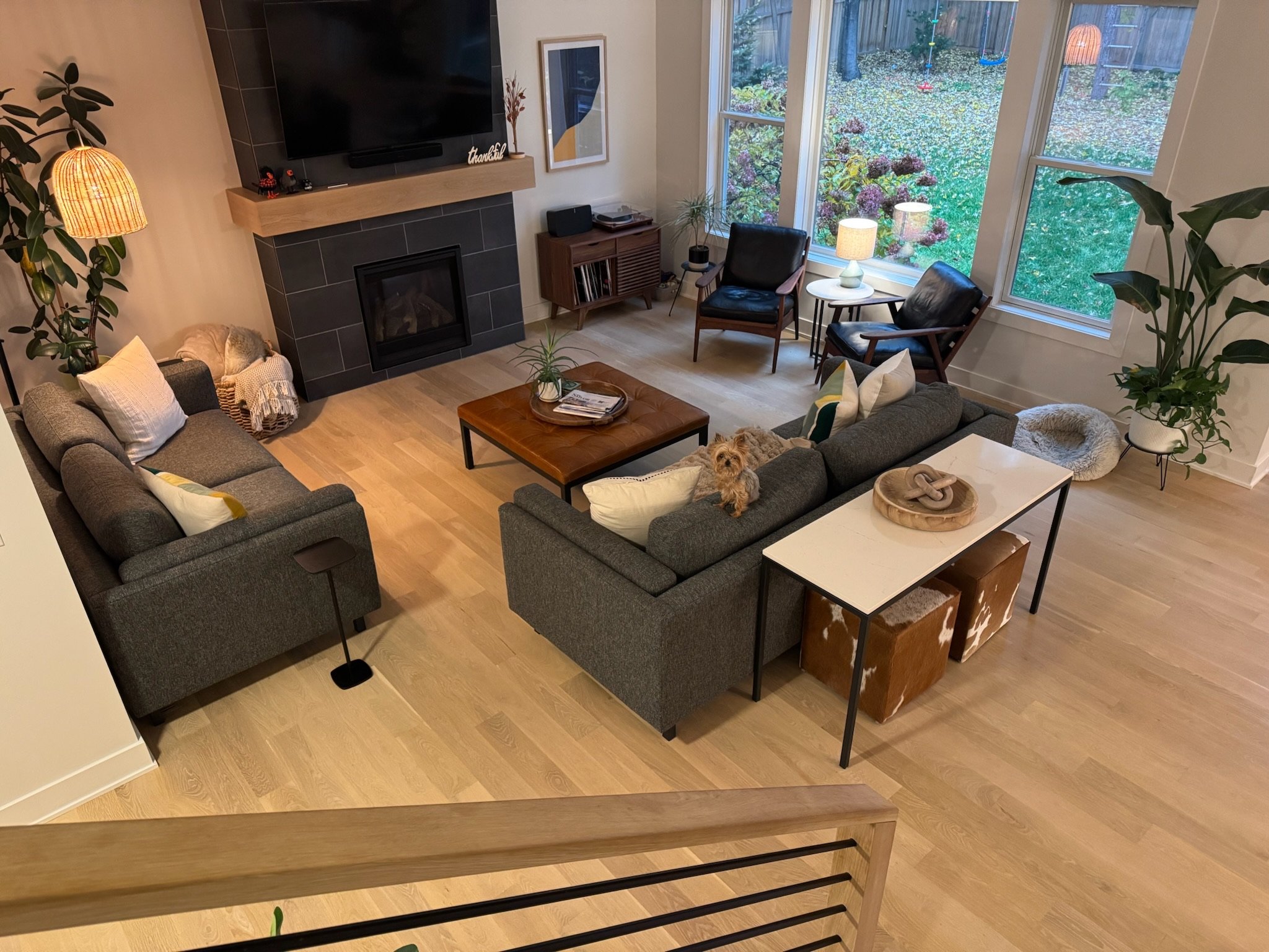 Living room with two dark gray sofas, a small dog on one sofa, wooden coffee table, two black armchairs near a large window, fireplace with mounted TV above, potted plants, lamps, and decorative items.