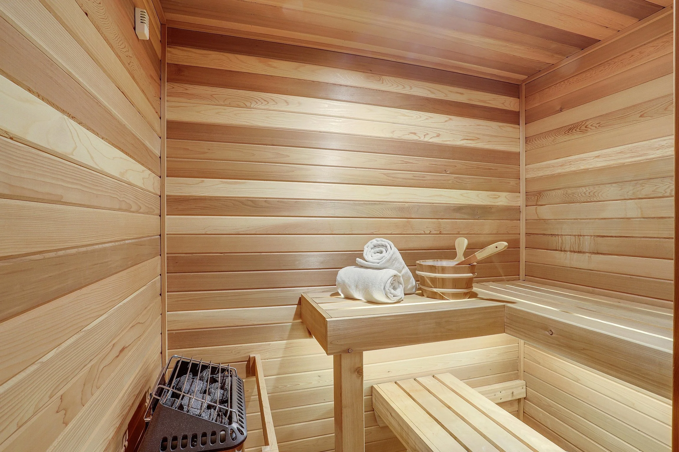 An interior of a wooden sauna with two rolled white towels, a wooden bucket, and a ladle on a wooden bench, and a heater with stones in the corner.