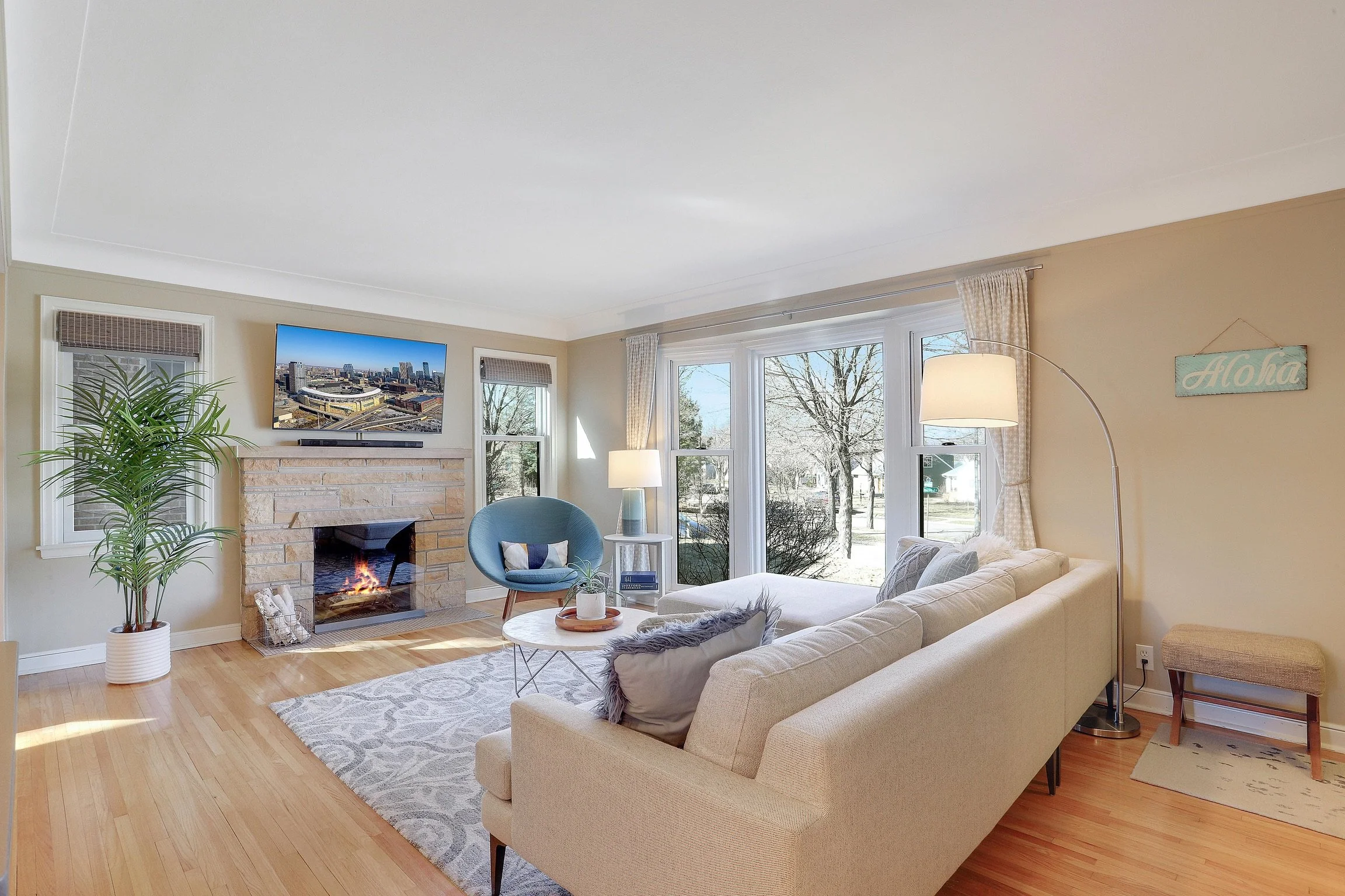 Living room with beige sectional sofa, white and gray pillows, wooden coffee table with white tray, floor lamp, blue accent chair, TV above fireplace, large windows with curtains, trees outside, house plant, and Aloha sign.
