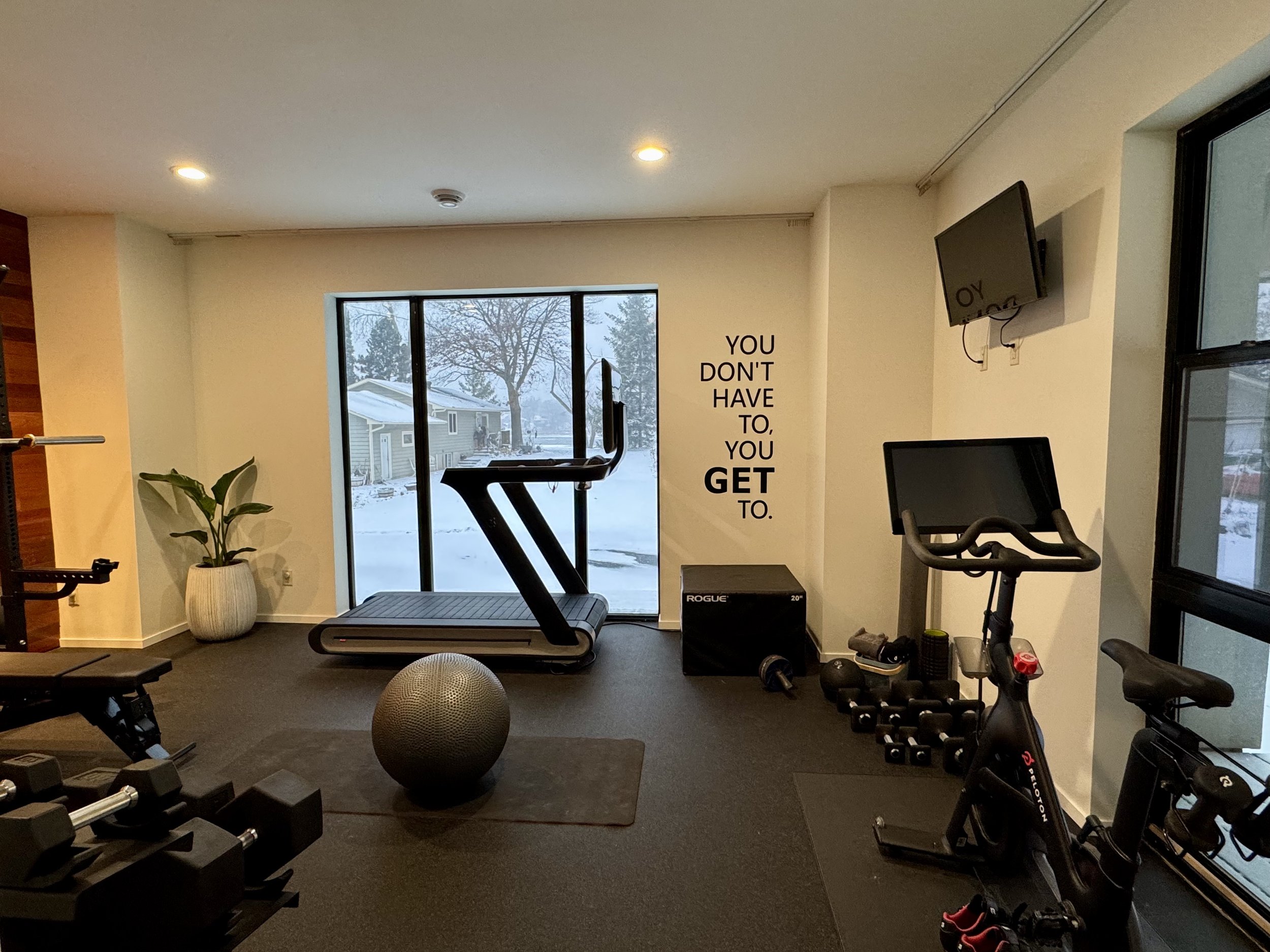 Home gym with treadmill facing large window showing snowy outdoor scene, wall decal saying 'You don't have to, you get to,' exercise bike, free weights, stability ball, potted plant, and wall-mounted TVs.