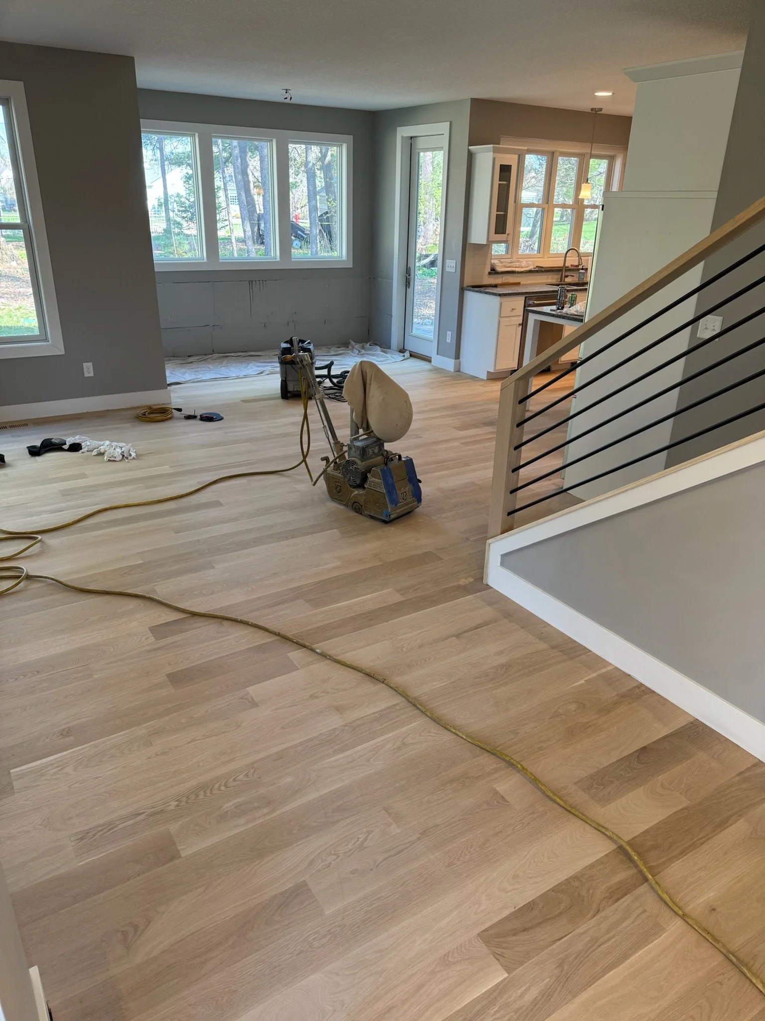 Living room with hardwood flooring, construction tools, and a large window overlooking a wooded yard, with an open kitchen in the background.