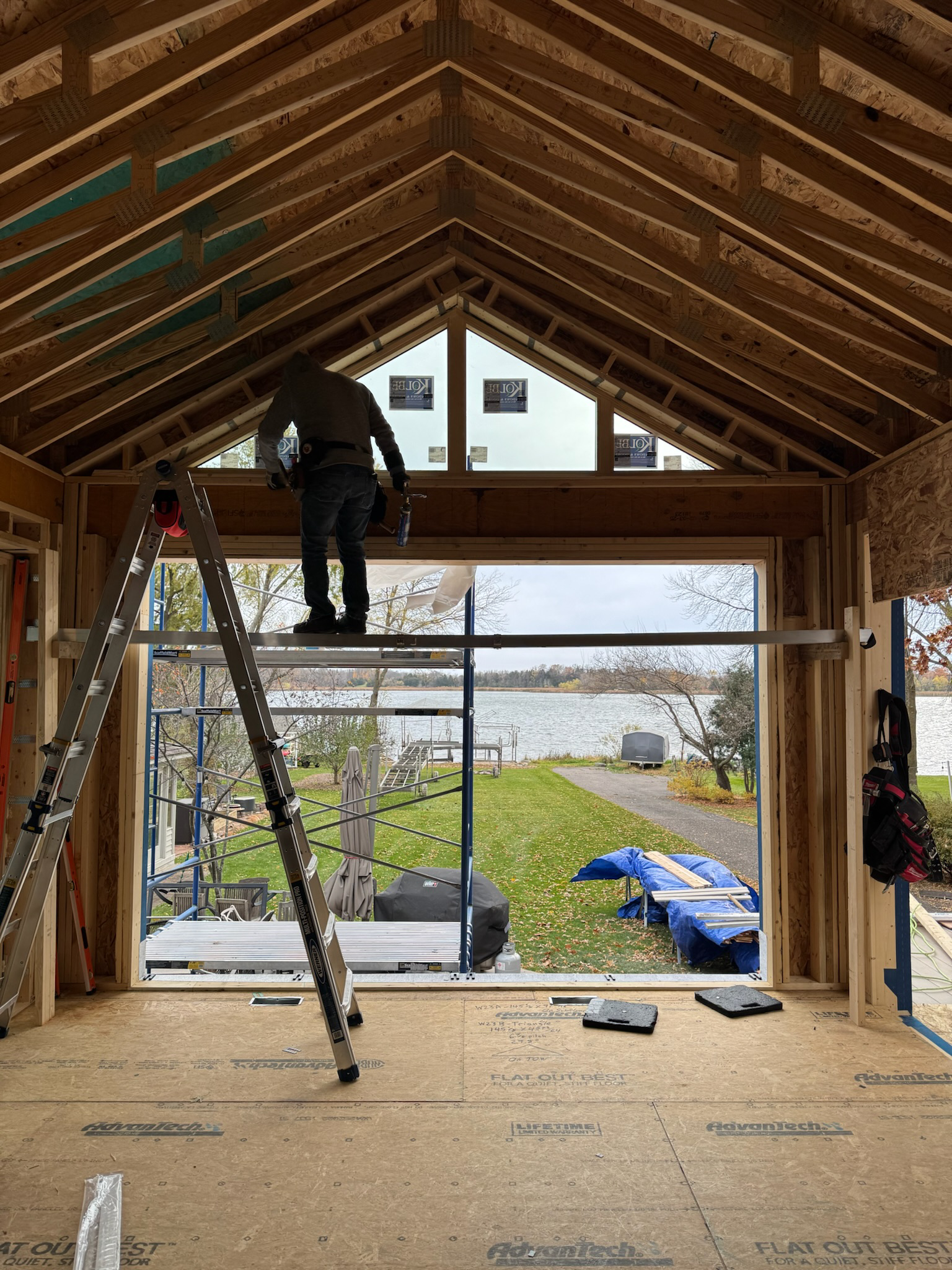 A person working on the interior framing of a house under construction, standing on scaffolding near a large opening with a view of a lake and outdoor scenery.