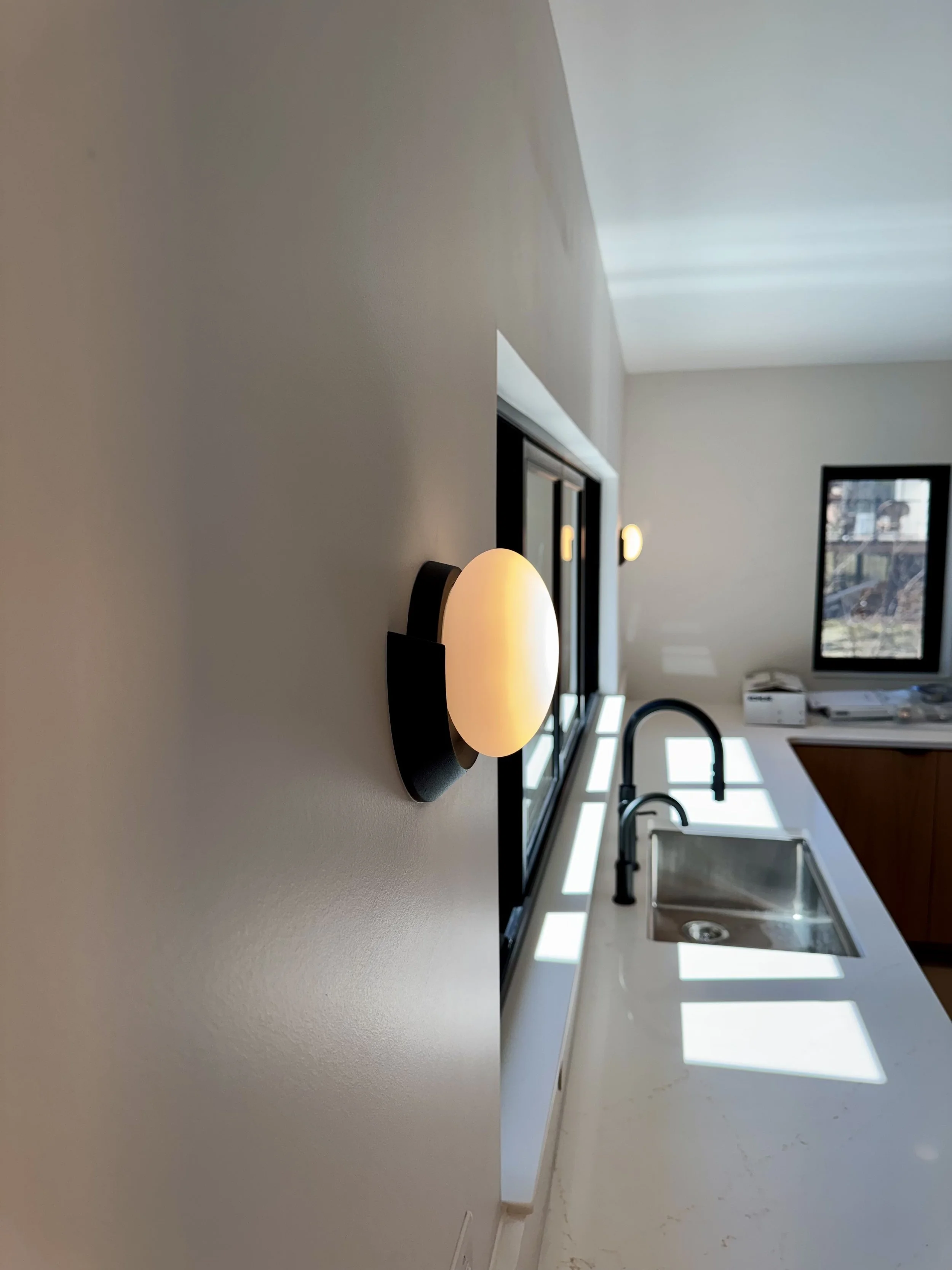 Modern kitchen with a wall-mounted light fixture, stainless steel sink, black faucet, white countertop, window with sunlight, and wooden cabinets.