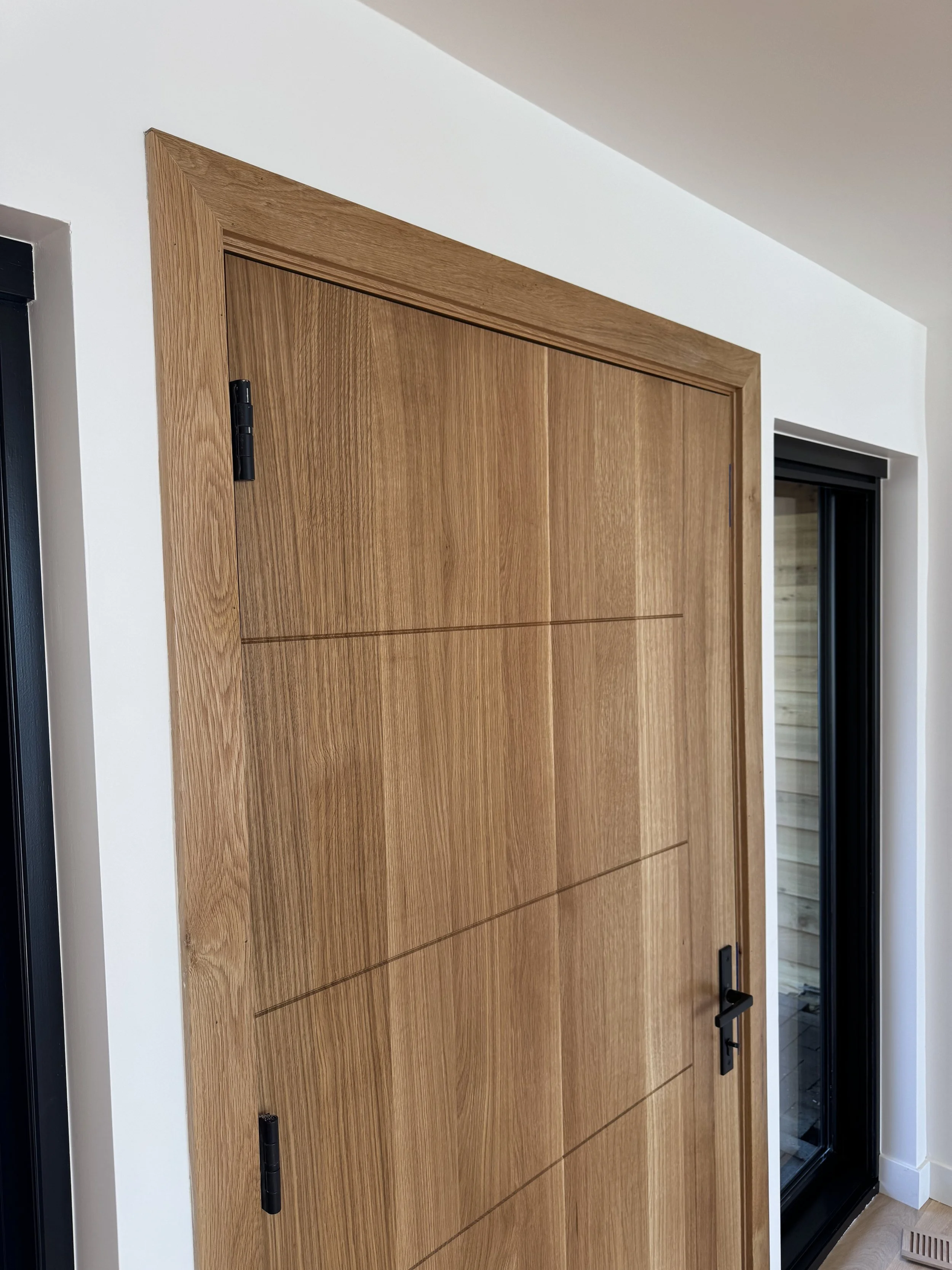 Wooden closet door with black hinges and handle, partially open, next to a window with black frame in a modern interior.