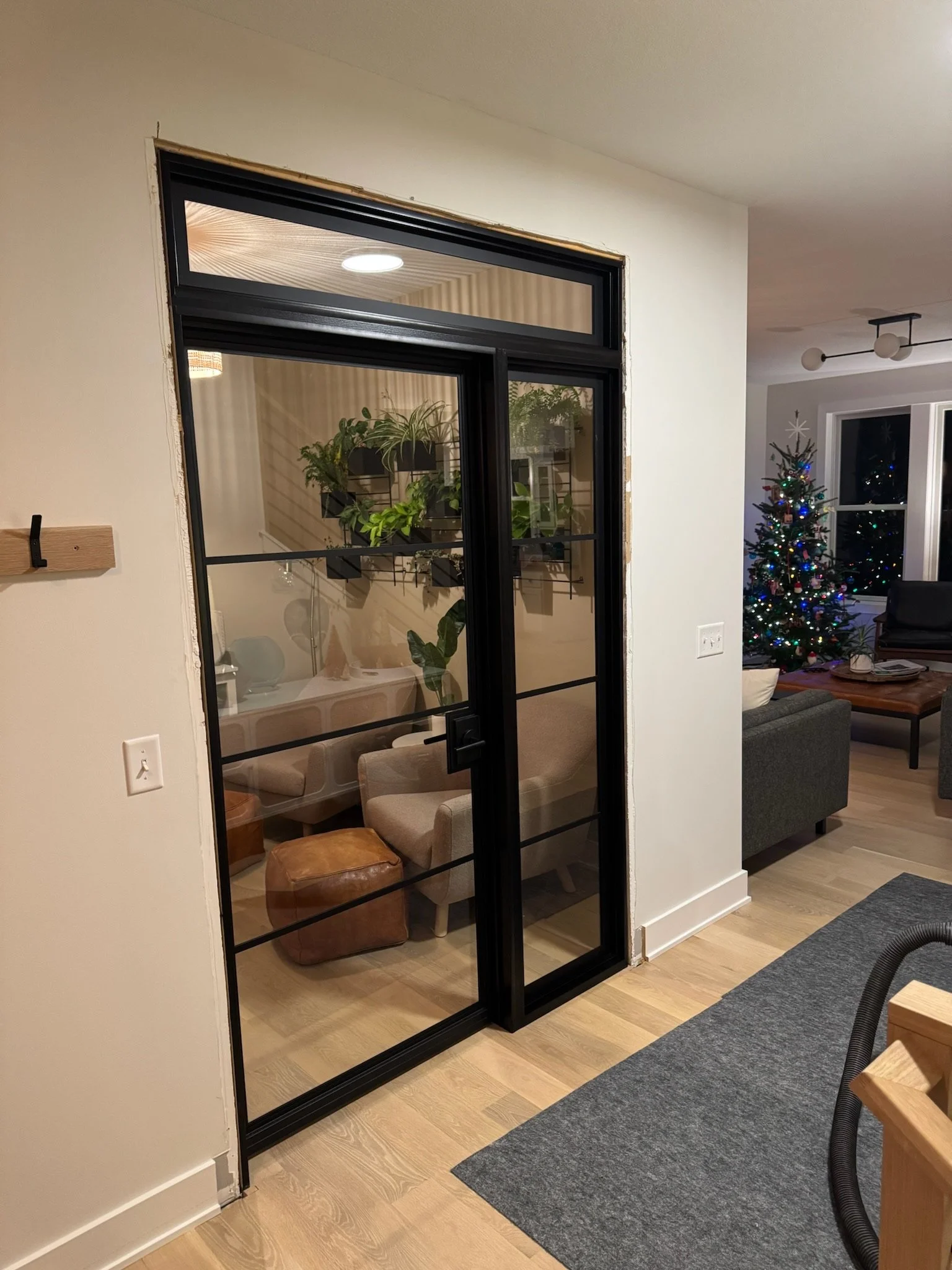 A black-framed glass door leading into a cozy room with a round table, beige chairs, and green houseplants on a wall shelf, with a living room visible in the background decorated for Christmas.