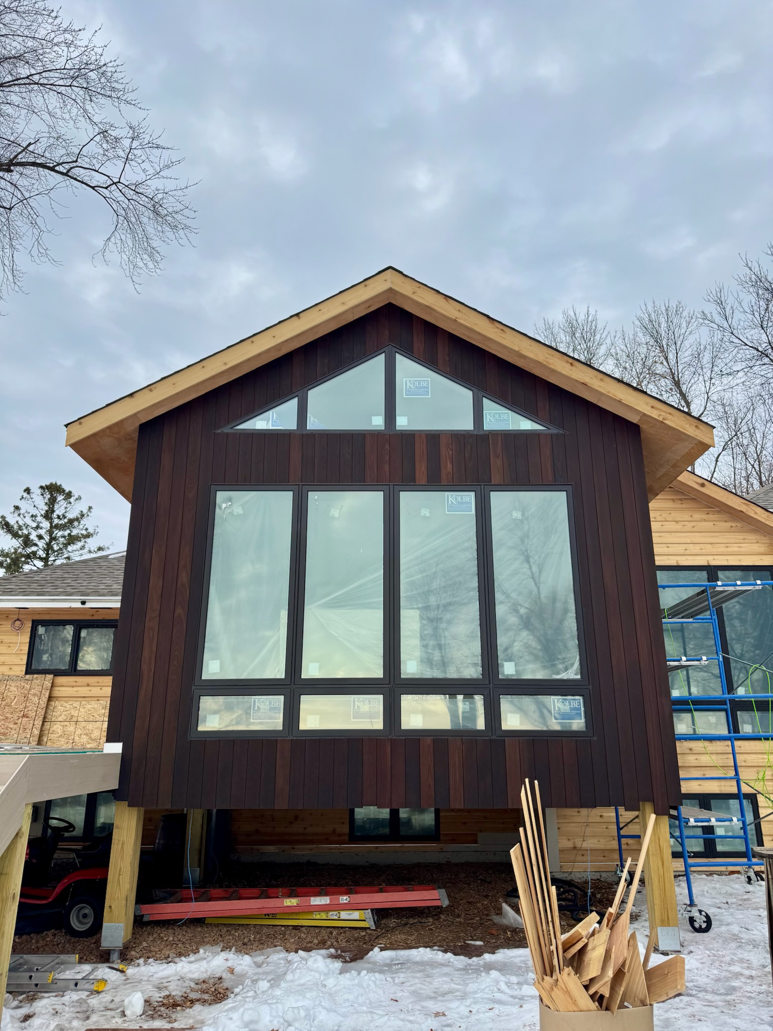 Under construction modern house with dark wood siding and large windows with a triangular roof, building materials and scaffolding visible, snow on the ground.