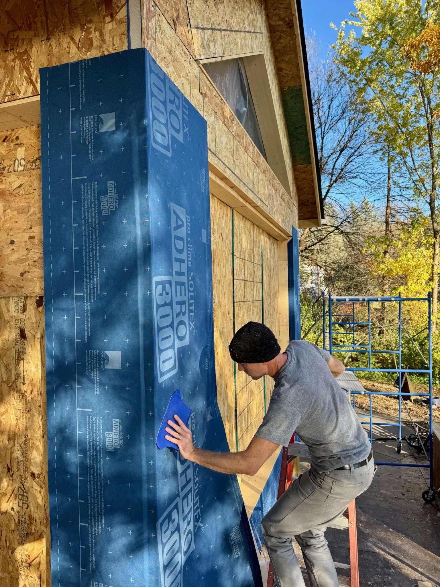 A man is installing blue house wrap on a wooden exterior wall under an open window, with a scaffold and trees in the background.