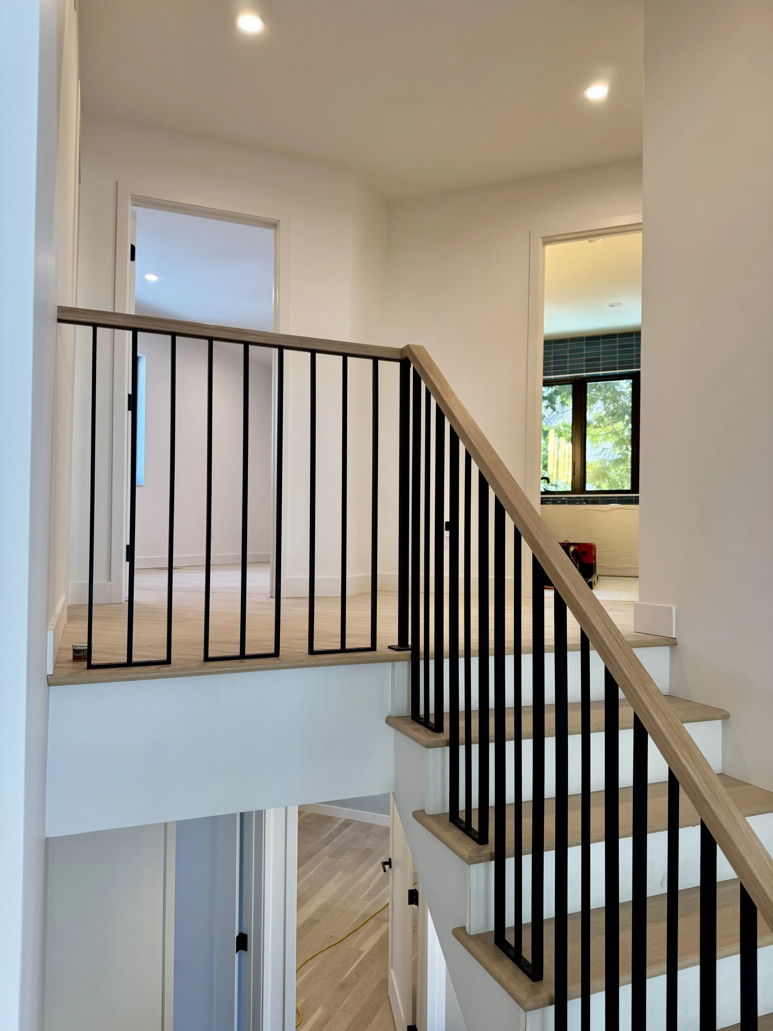 Interior view of a staircase with black metal railings and wooden handrails, leading to an upper floor with white walls, a ceiling with recessed lighting, and visible doorways to other rooms.