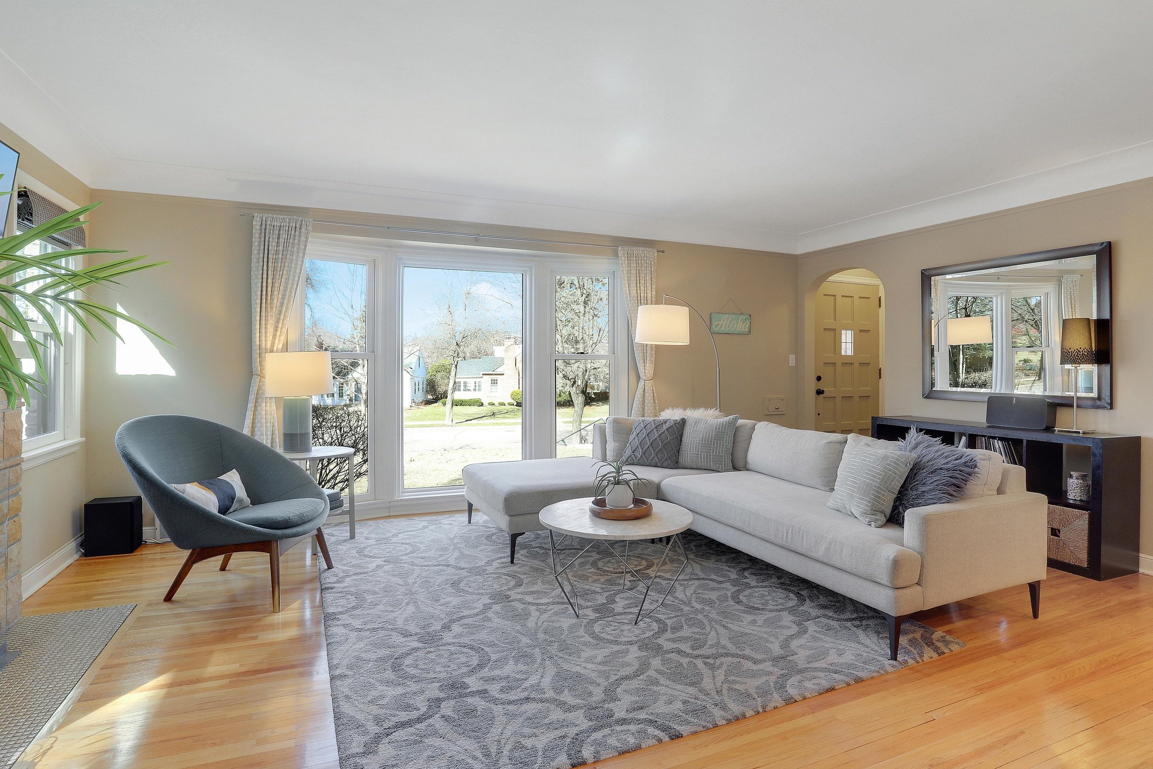 Bright living room with large window, white sofa with pillows, gray armchair, coffee table with plant, mirror on wall, and lamp next to sofa, overlooking a yard.