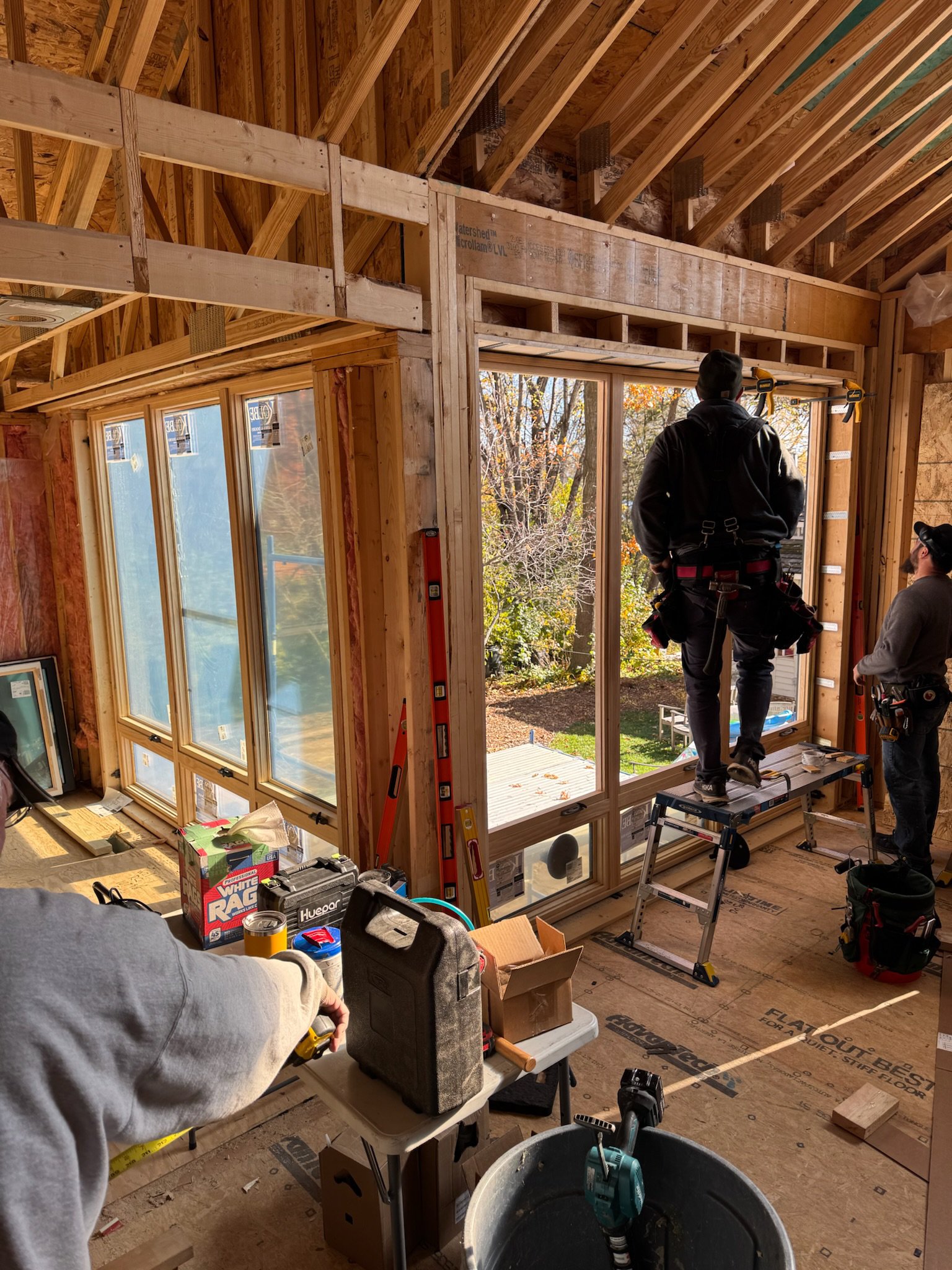 Construction workers installing large window panels in a wooden house under construction, with tools and materials around.