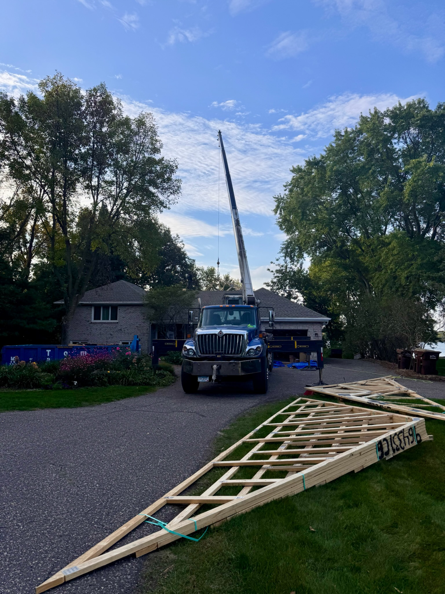 A crane truck parked on a driveway near a house during daylight, with a toppled wooden structure lying on the grass in front of it, surrounded by trees and landscaping.
