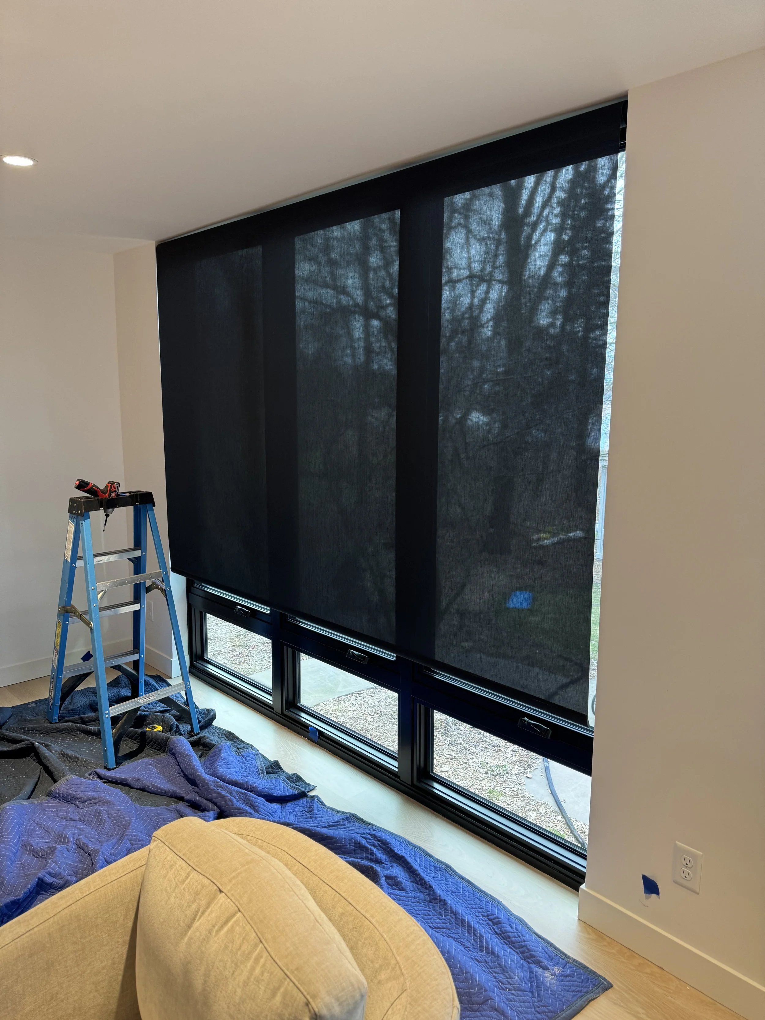 Interior of a room with large windows covered by black window shades, a blue ladder, and a yellow ottoman.