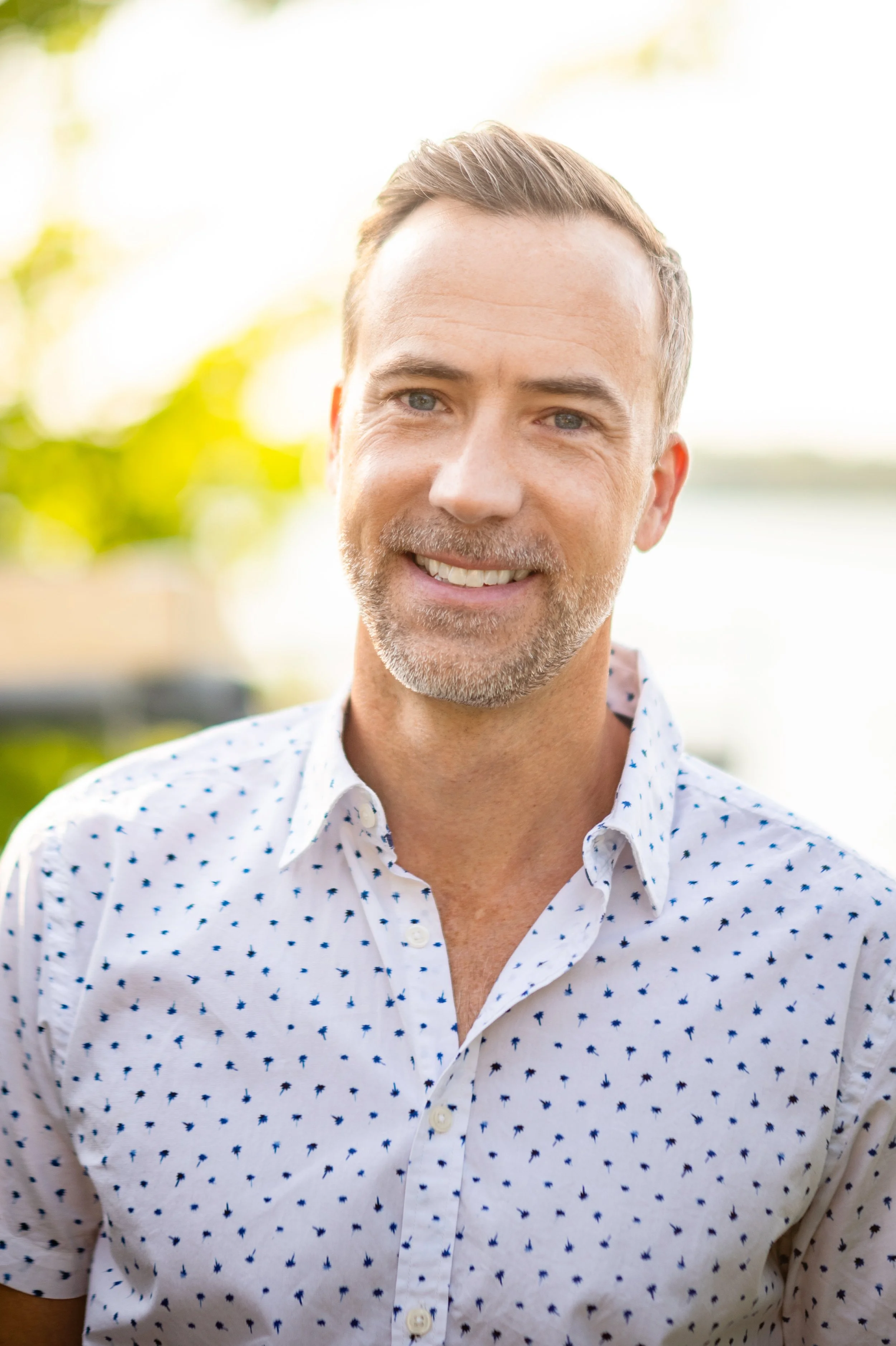 Smiling middle-aged man with short brown hair and a beard, wearing a white shirt with small dark blue patterns, outdoors with blurred green foliage in the background.