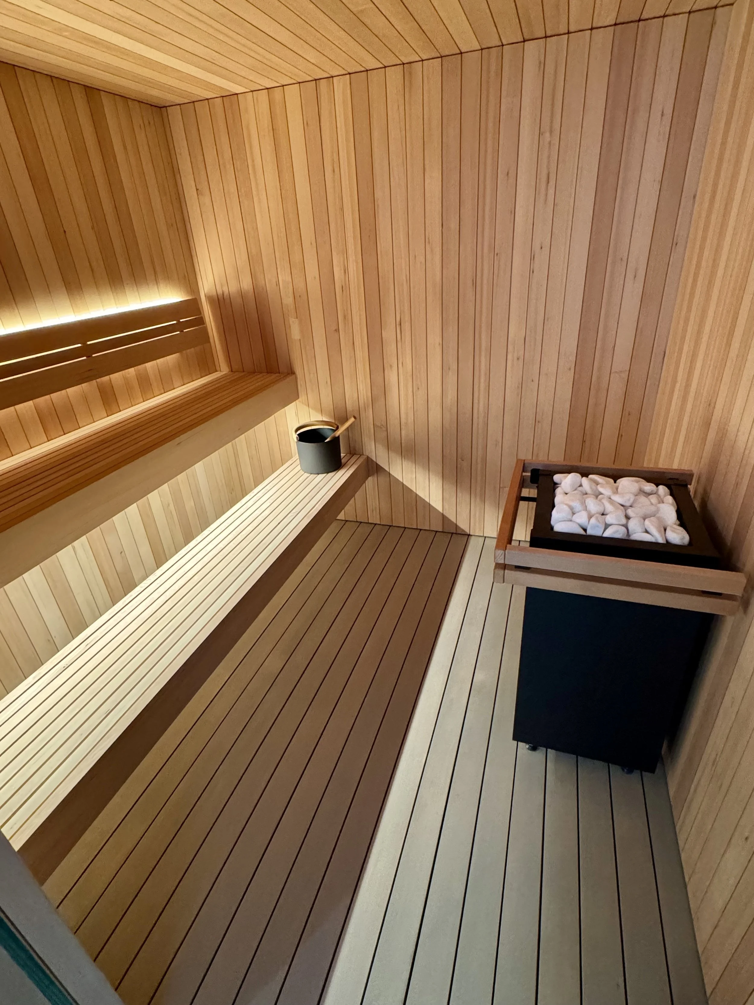 Interior of a wooden sauna with benches, a bucket with a ladle, and a heater filled with white stones.