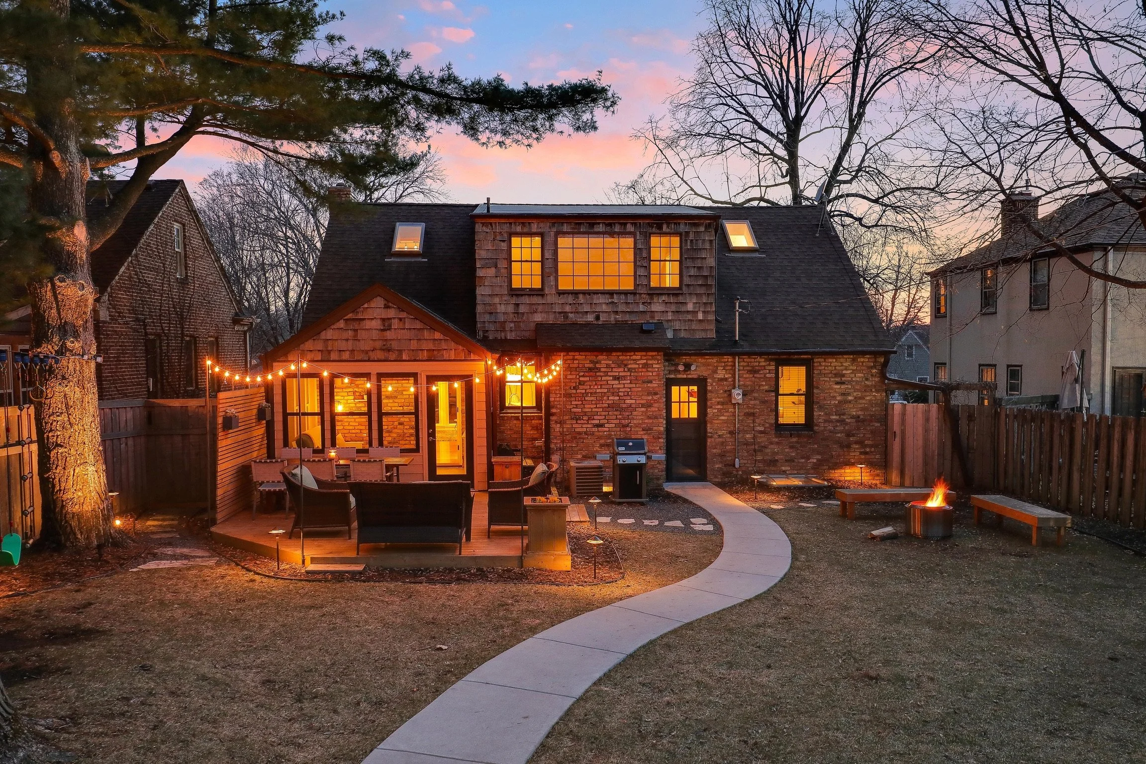 The backyard of a brick house with a deck, string lights, and a fire pit, during sunset.