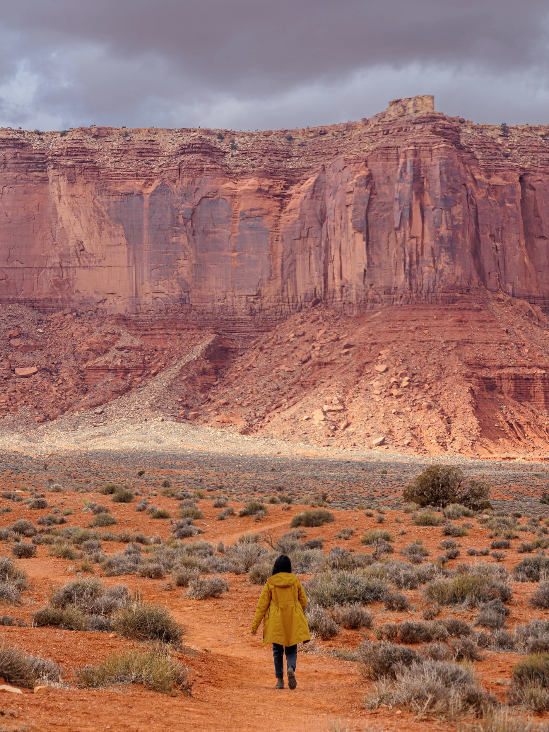 Person in a yellow jacket walking on a dirt trail through a desert landscape with red rocks and cliffs, under a cloudy sky.