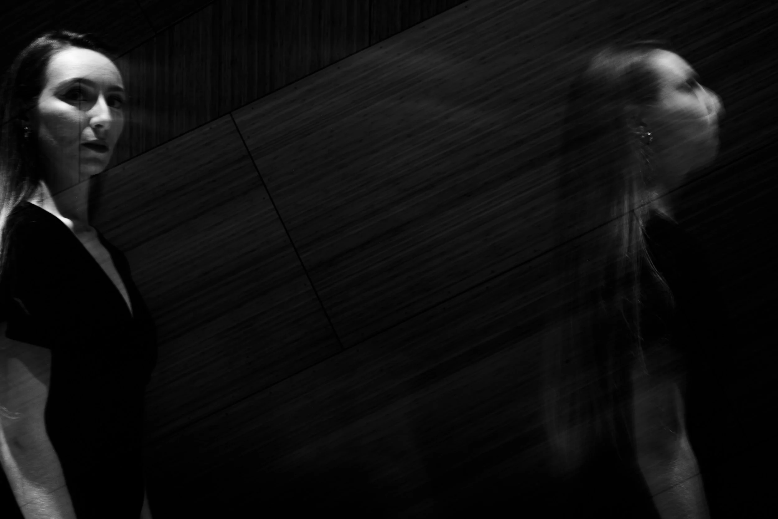 A black and white photo of a woman looking at her reflection in a mirror with a wooden background. The reflection appears blurred due to movement.