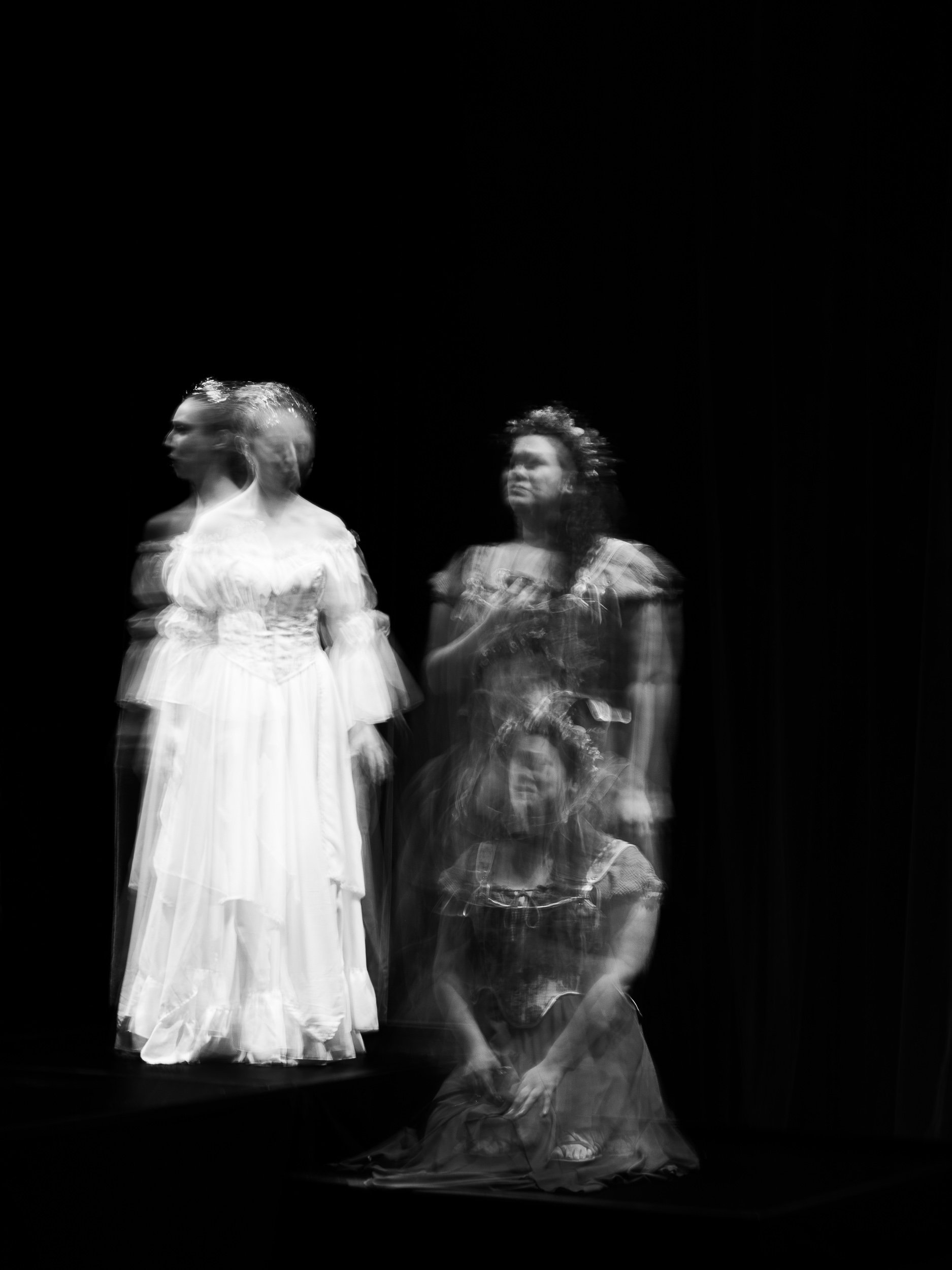 Black and white photo of three women dressed in vintage-style dresses, with their images appearing blurred or duplicated, against a dark background.