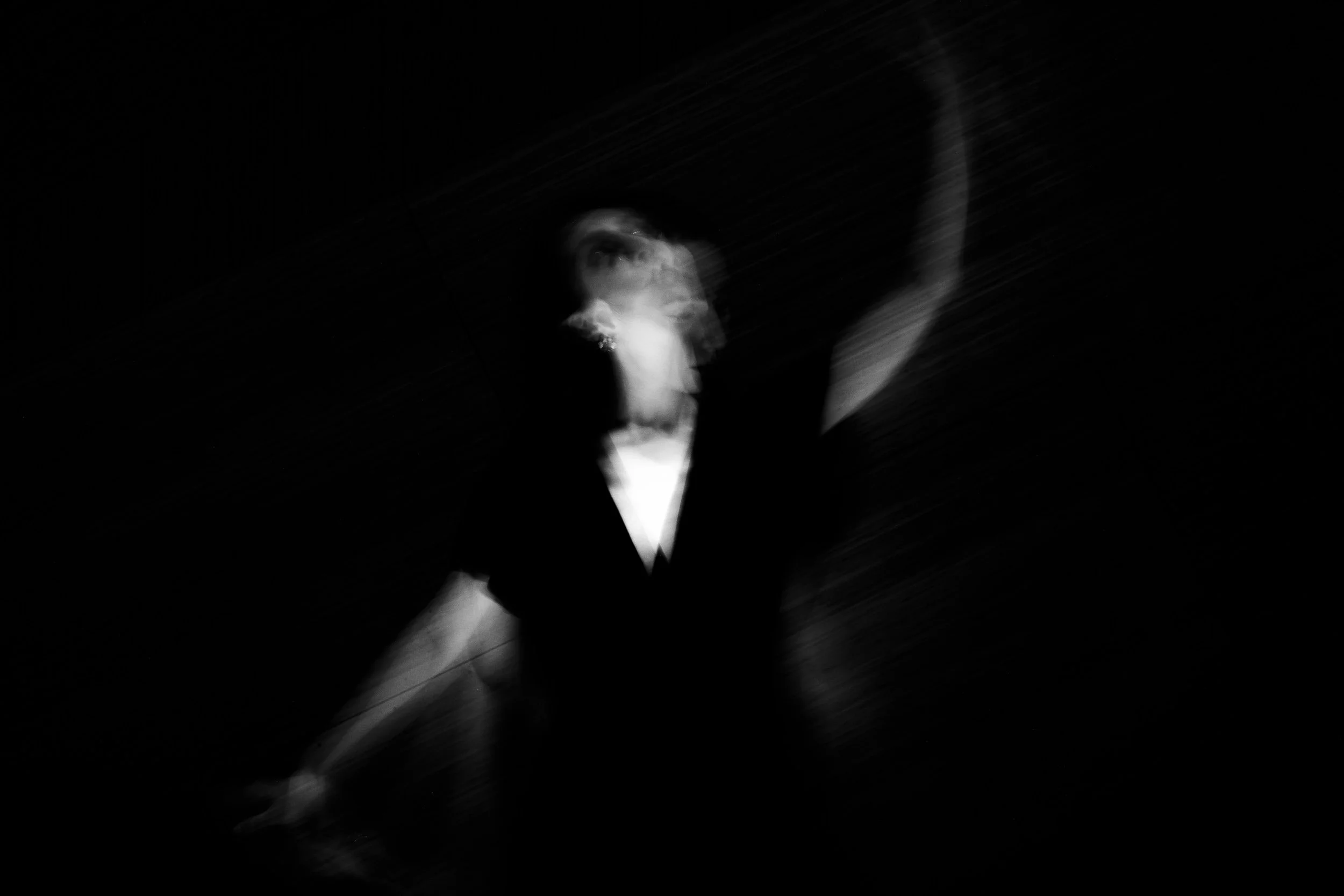 Black and white photo of a woman raising her arm with motion blur, with her face partially visible and looking upward, in a dark background
