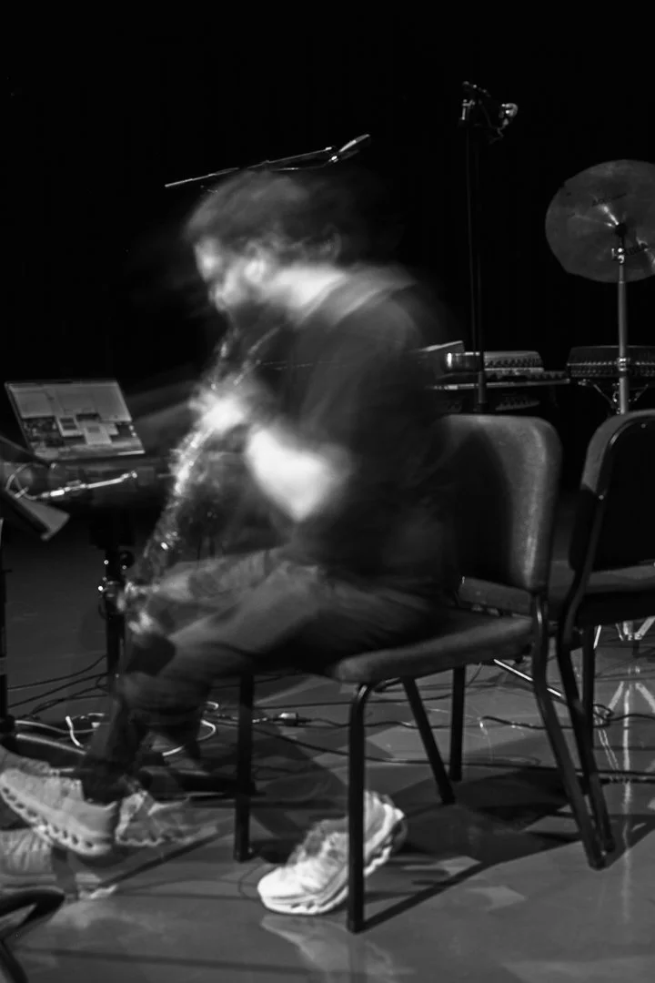 A person sitting in a chair on a dimly lit stage, blurry motion indicating movement, surrounded by musical equipment including a laptop, microphone, and cymbal.