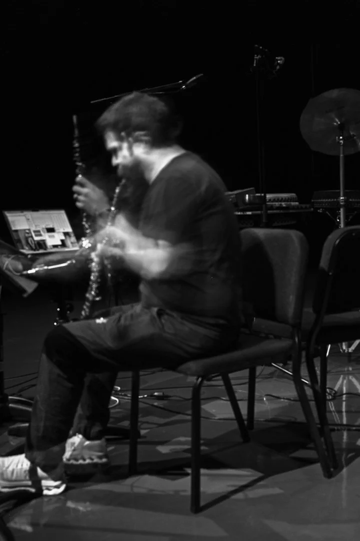 A person playing a saxophone on a dimly lit stage, with musical equipment and a cymbal visible in the background, captured in black and white with motion blur.