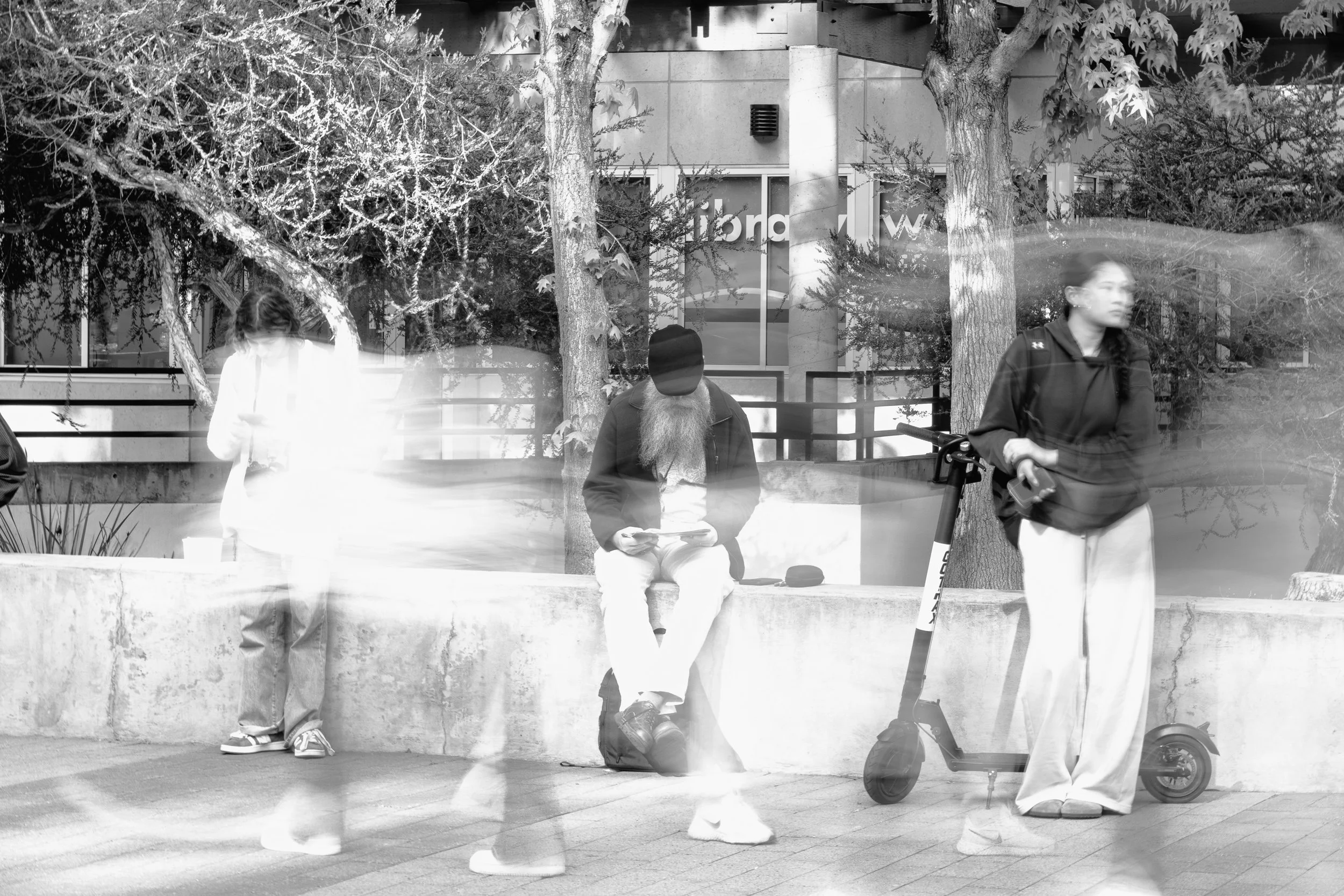 Three people sitting and standing on a sidewalk in a city park with trees in the background; a person with a long beard reading a book, a woman using a scooter, and another woman looking at her phone.