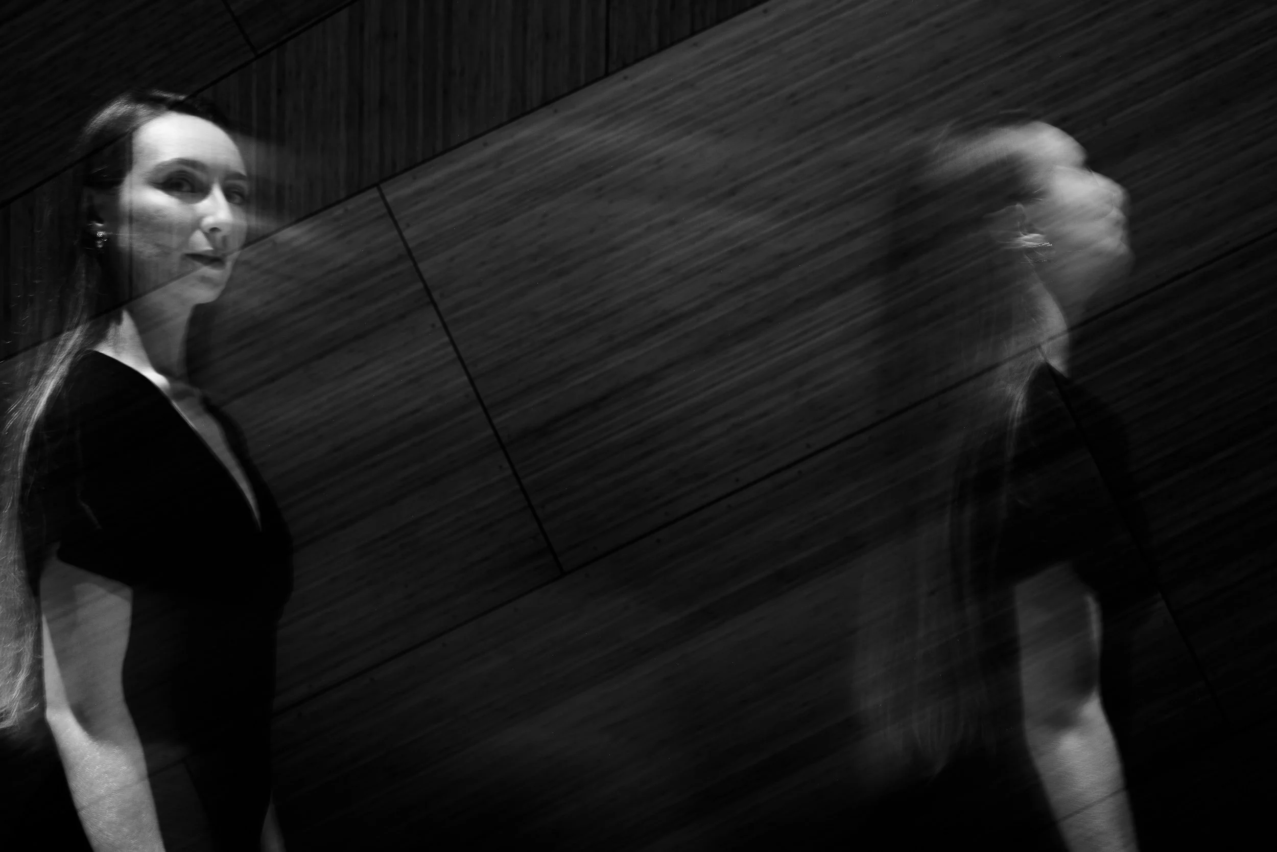 Black and white photo of a woman with dark hair, reflected in a mirror, with a blurred second reflection of her in motion, against a wooden wall background.