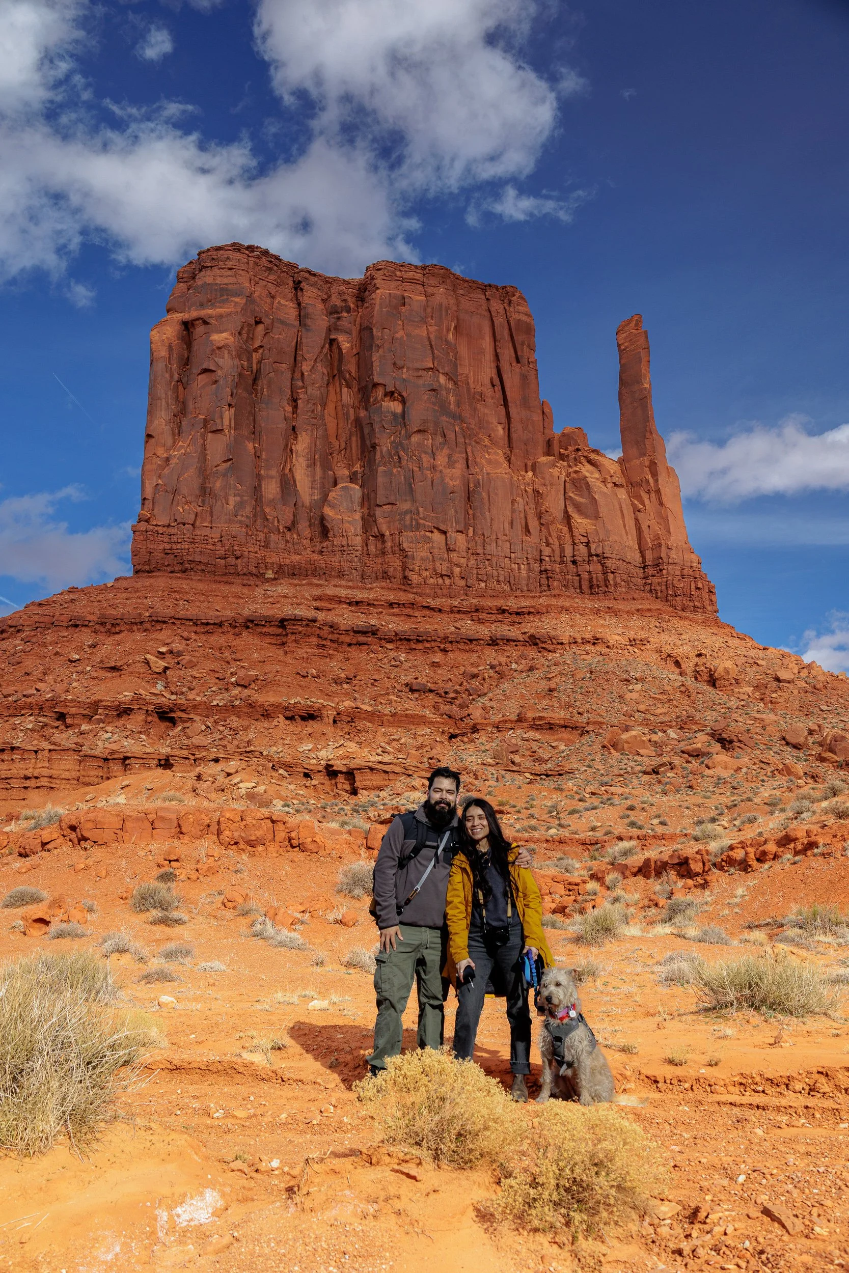 Two hikers, a man and a woman, standing with their dog in a desert landscape with a large red rock formation and a partly cloudy sky in the background.