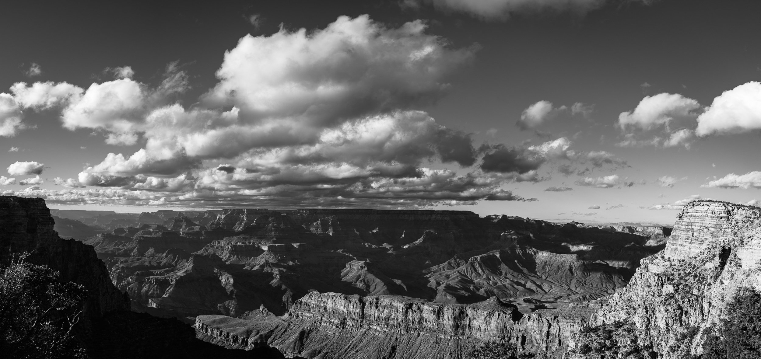 Black and white photo of the Grand Canyon showing layered rock formations, cliffs, and a partly cloudy sky.