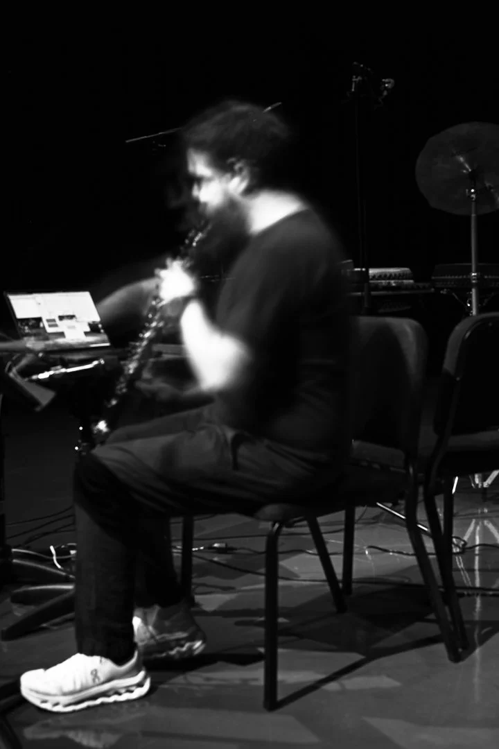 A person sitting on a chair playing a clarinet in a recording studio or stage, with a laptop and musical equipment around.