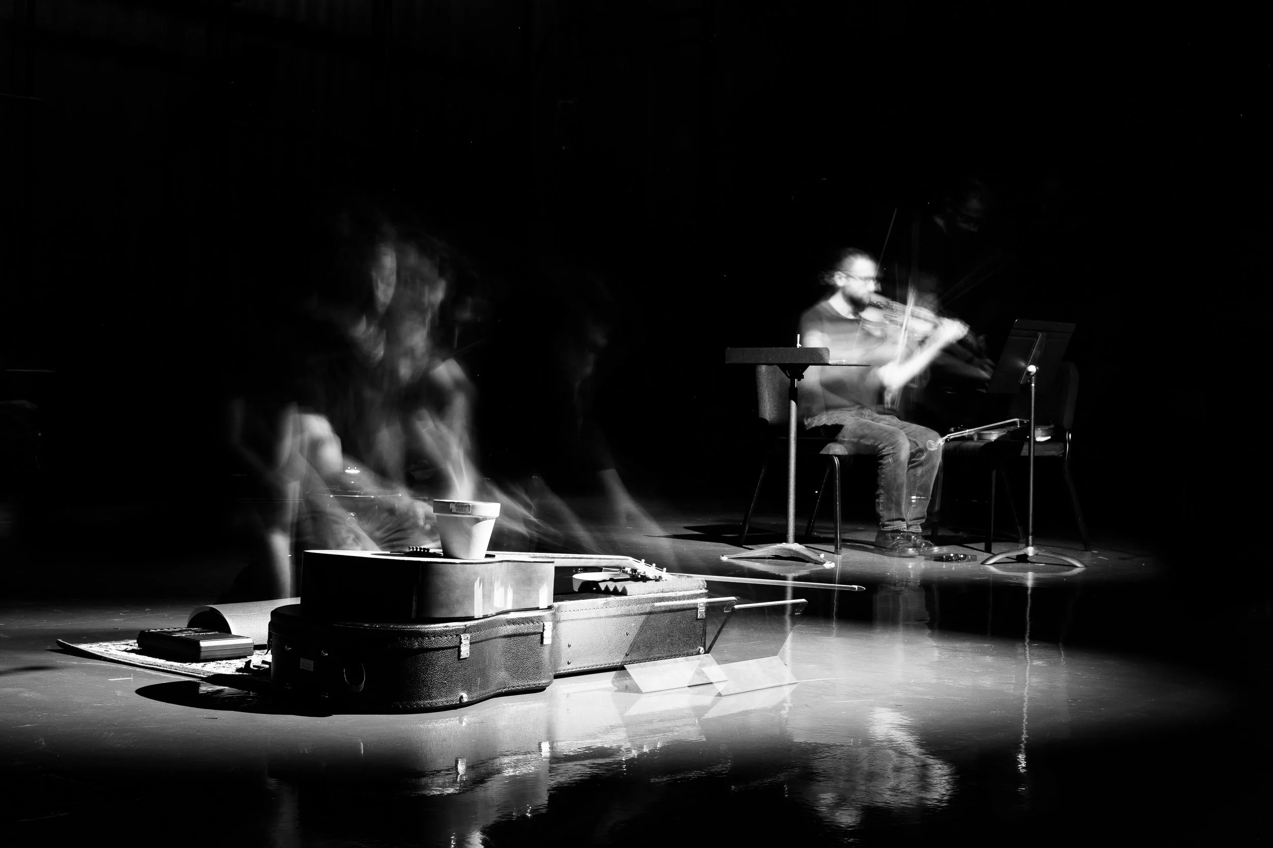Black and white photo of a live musical performance with a person playing violin, one sitting on a chair, on a dark stage with minimal lighting, creating a motion blur effect.