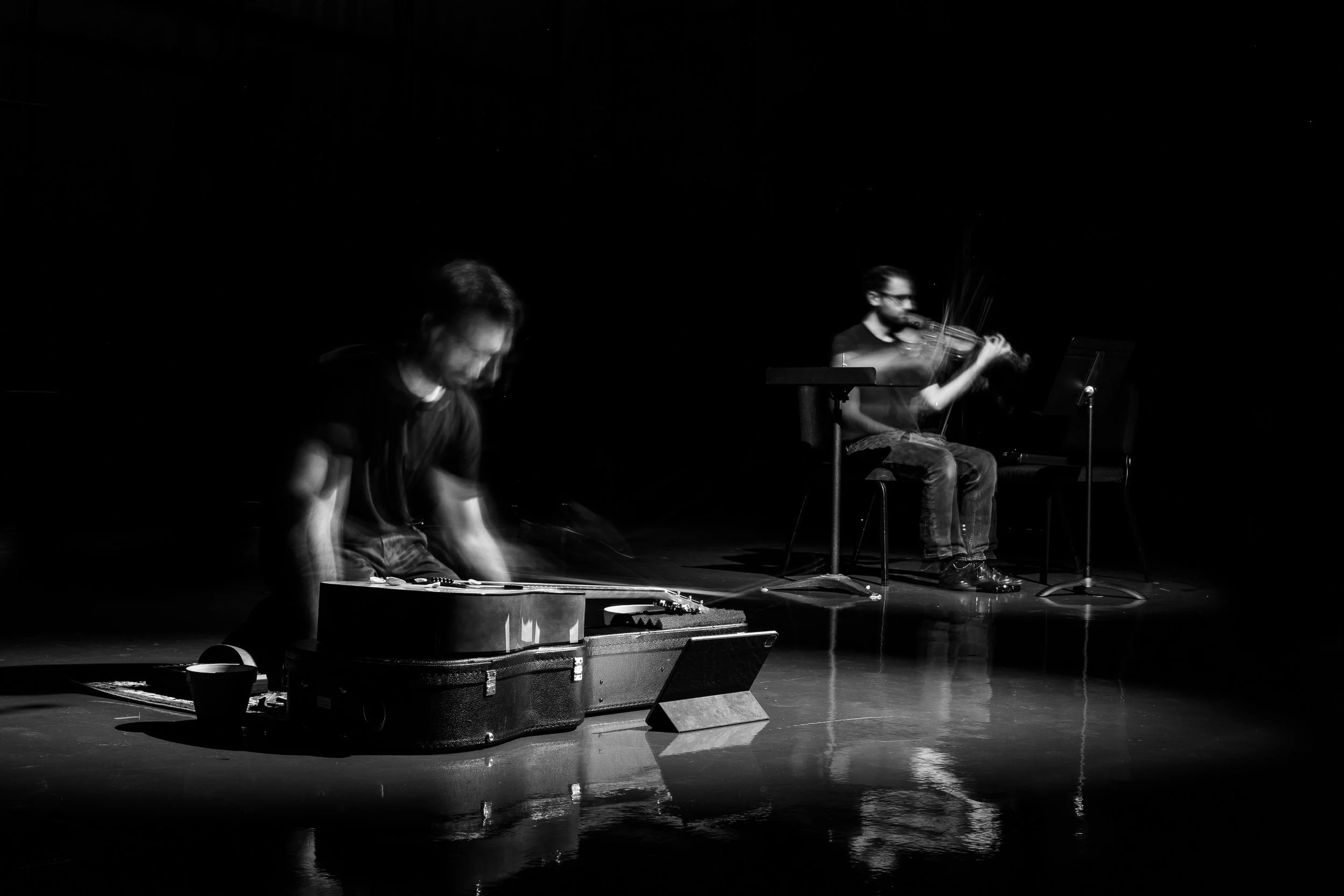 Two musicians performing on stage: one playing a guitar and the other playing a violin, both captured with motion blur in a dark setting.